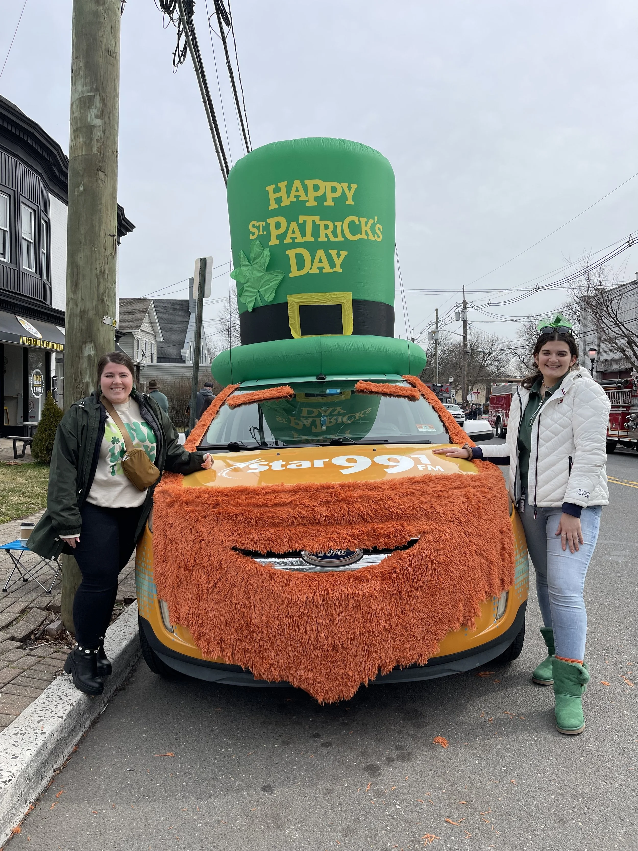 Two women standing beside a decorated yellow car with an orange furry cover on the hood and a large inflatable green hat on top that says 'Happy St. Patrick's Day'. The women are smiling, one wearing a white jacket and green boots, the other in a dar