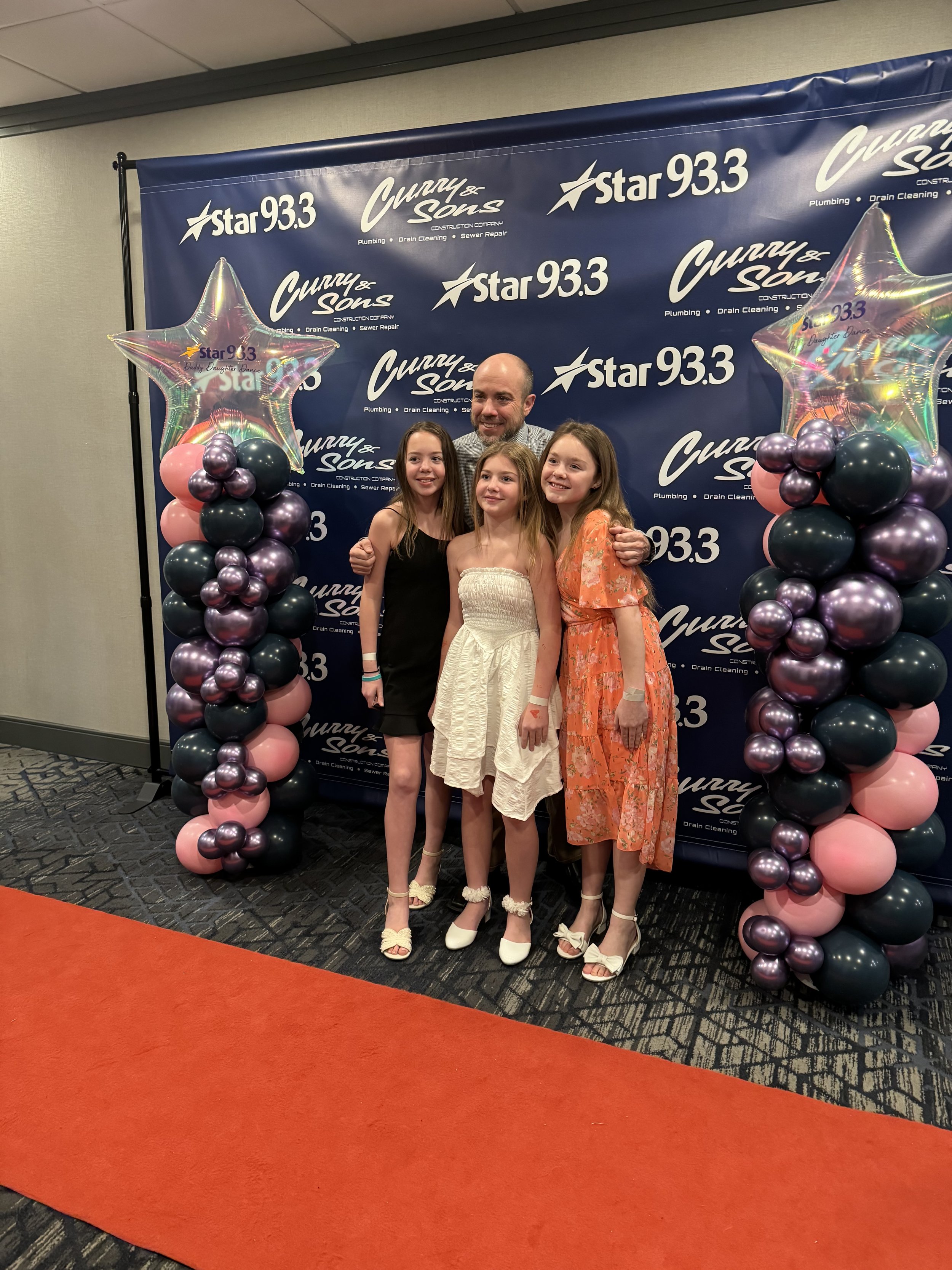 A group of four people, three young girls and one man, standing together in front of a blue backdrop with the logos 'Star 93.3' and 'Curry & Sons'. The backdrop features large star-shaped balloons with pink, purple, and metallic colors, decorated wit