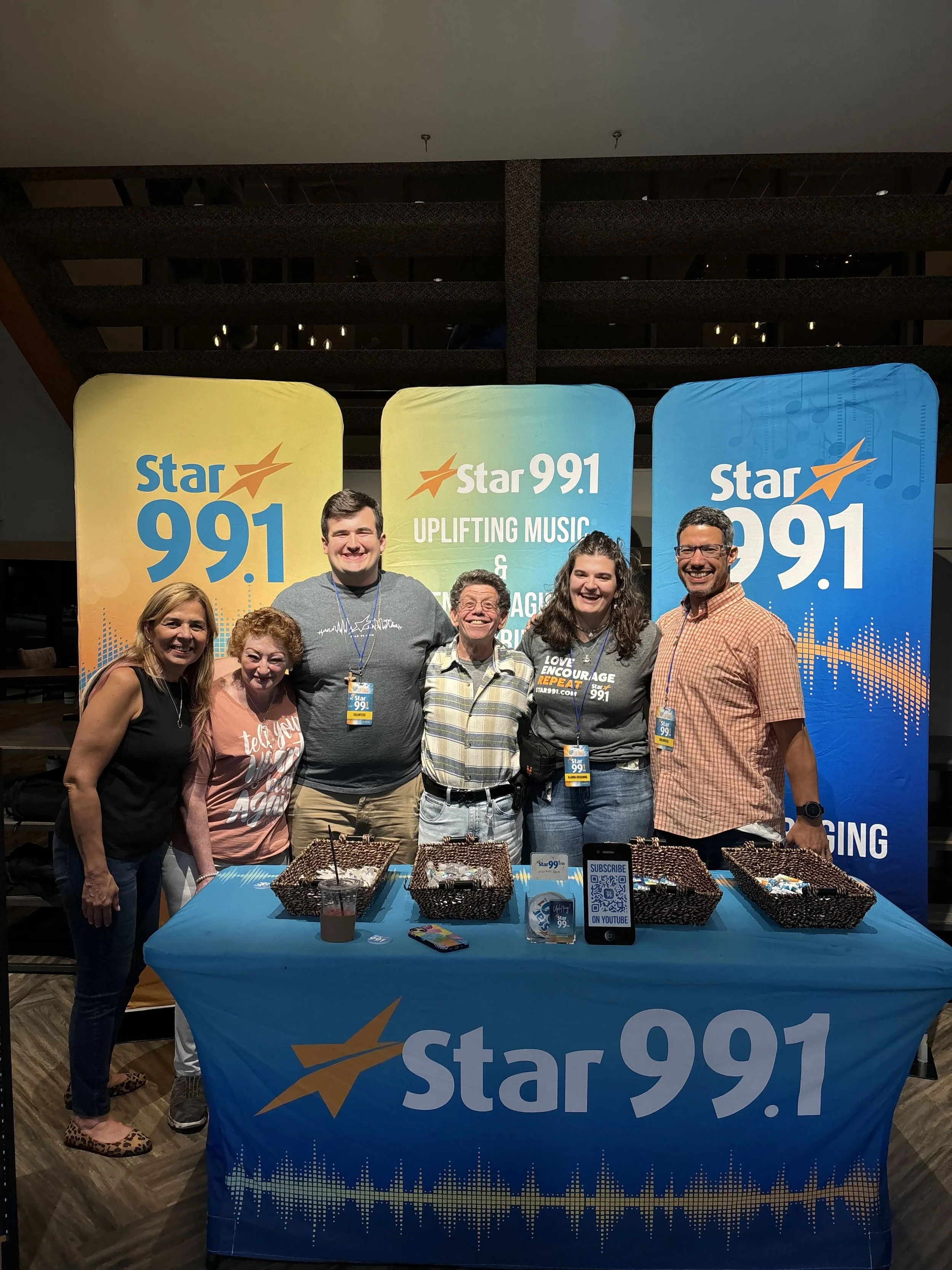 Group of six people standing behind a table with baskets at a Star 99.1 radio station promotional event.