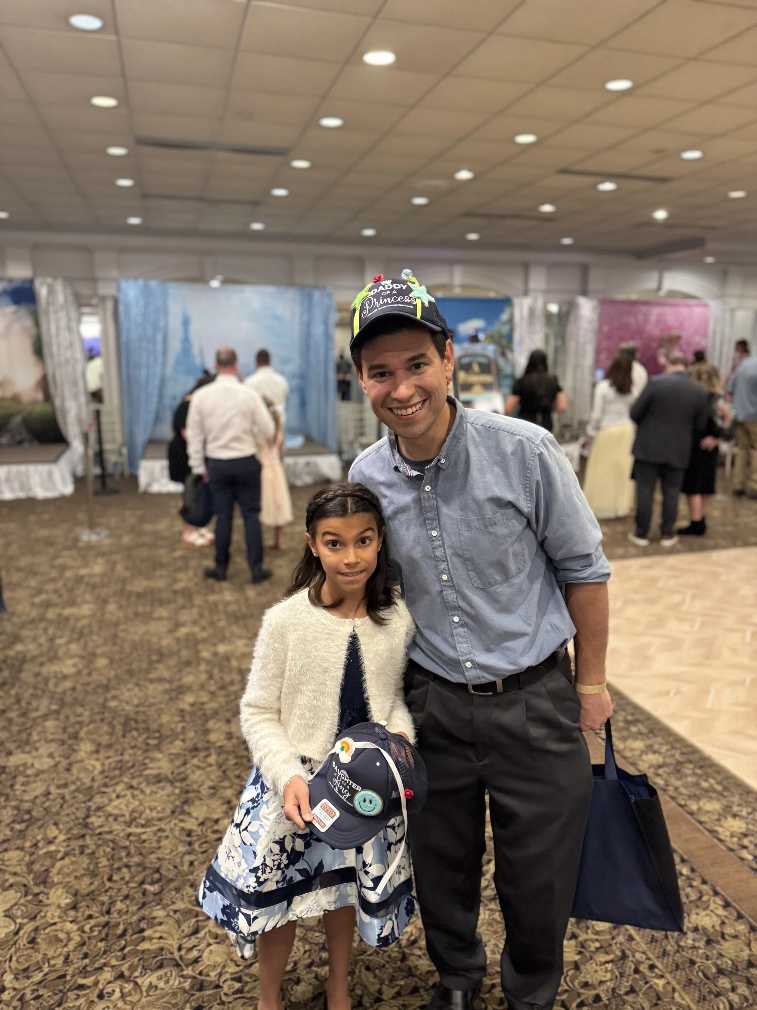 A young girl and a man are smiling and posing together in a decorated indoor venue, possibly a birthday celebration, with other people and decorated booths visible in the background.