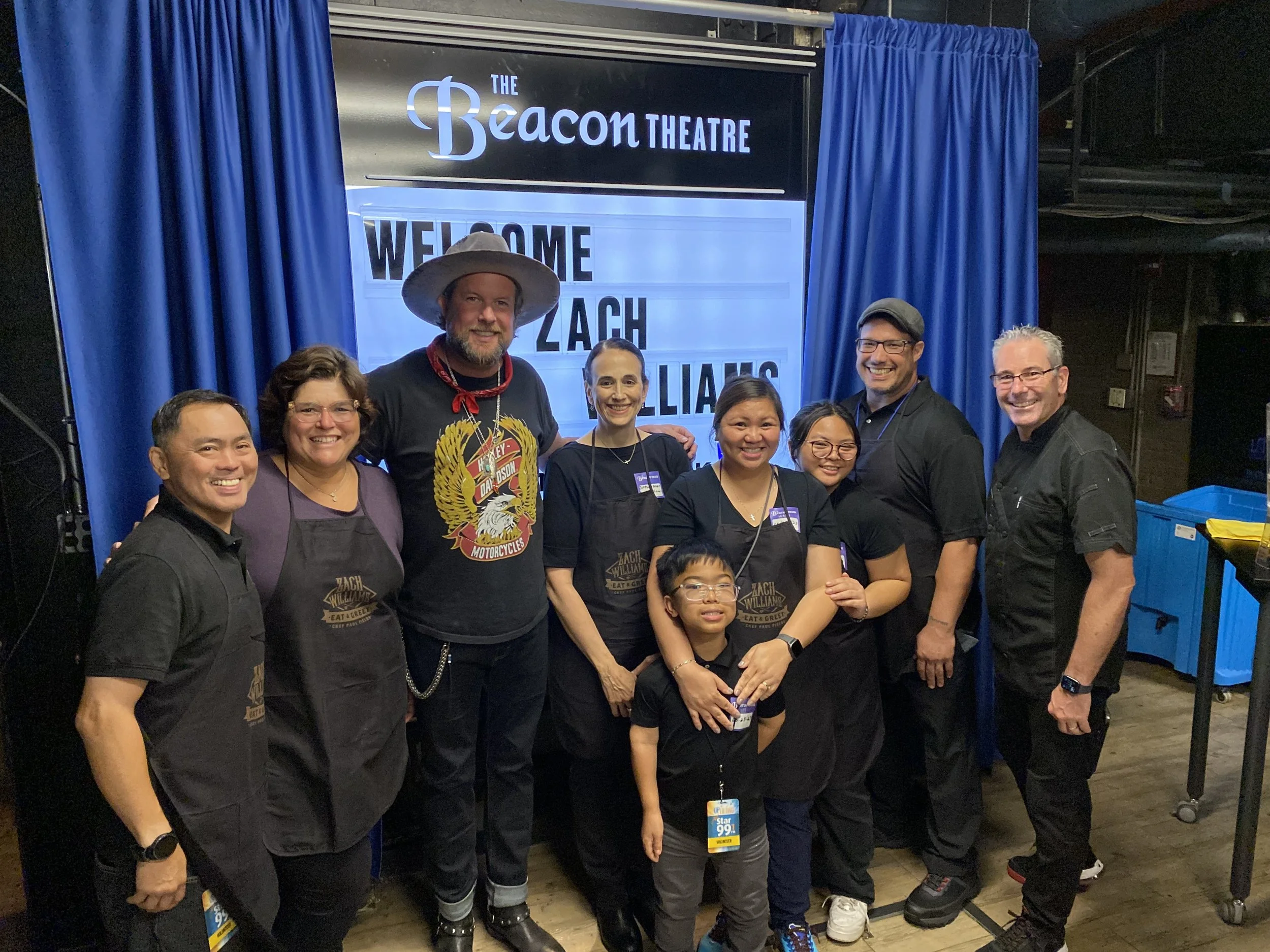 Group of nine people posing in front of a blue curtain and a sign that says 'The Beacon Theatre' and 'Welcome Zach Williams.'