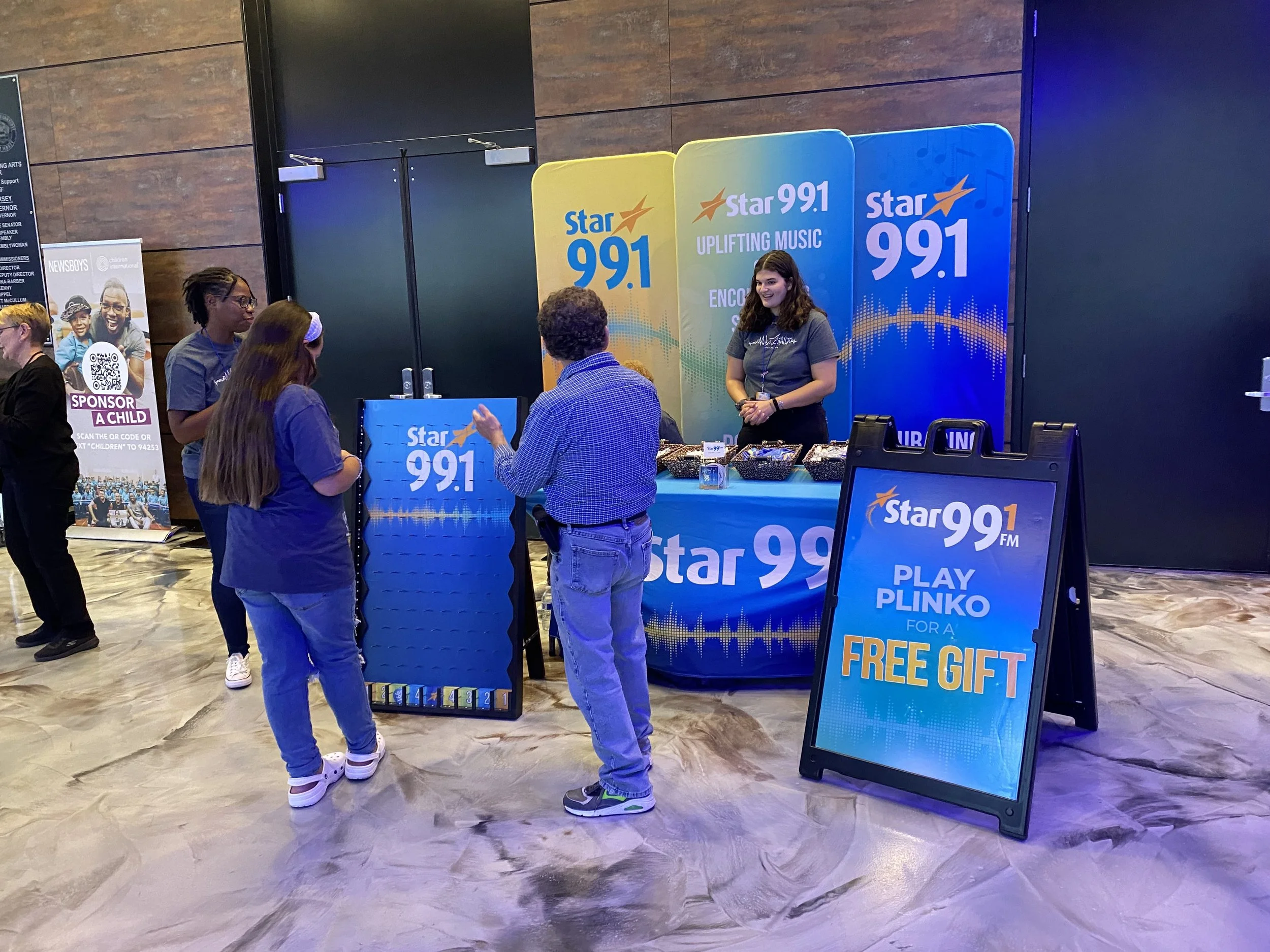 People gathered at a promotional booth for Star 99.1 FM, which includes a display board and a sign offering a free gift for playing Plinko. The booth staff is engaging with attendees, and there is a large colorful backdrop with the radio station's br