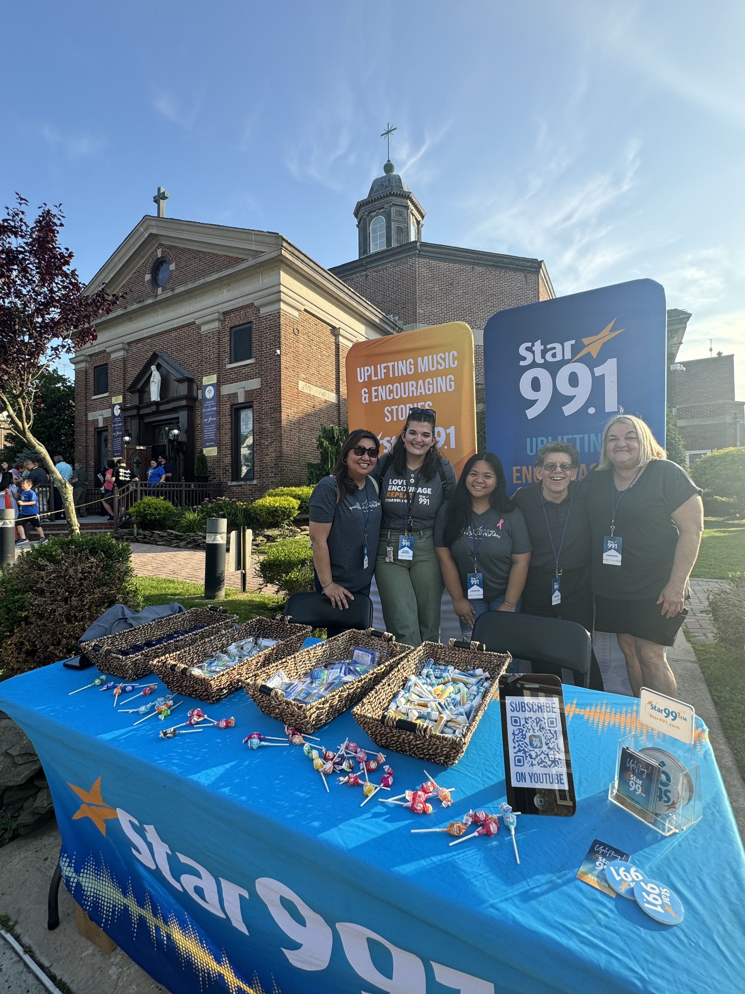 Group of five women standing behind a table with promotional items at an outdoor event in front of a brick church. The table displays candy and hand sanitizer; signs promote Star 99.1 radio station and encouraging messages.