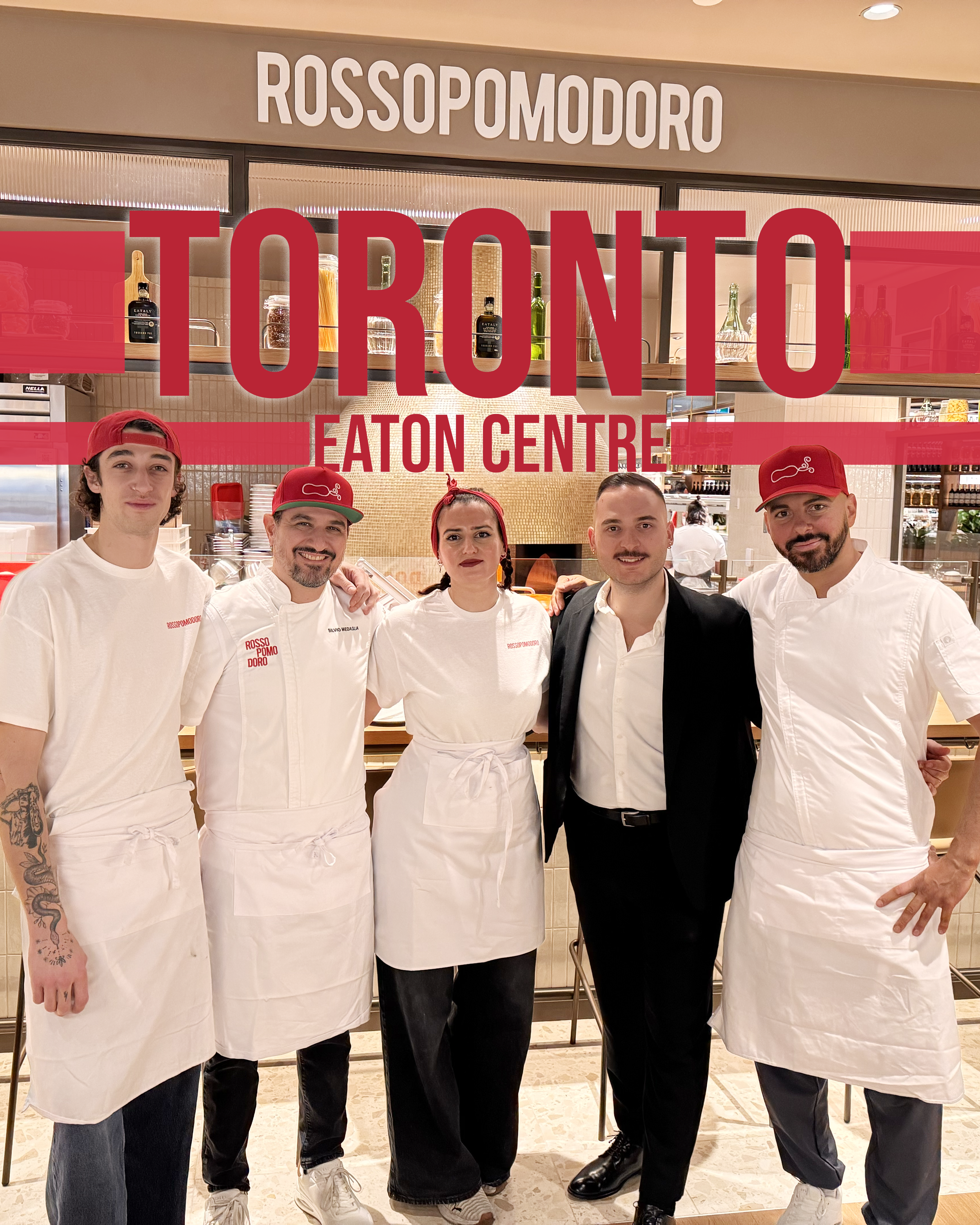 Group of five people standing in a restaurant kitchen, three are dressed in white chef uniforms and two in black suits, with red hats, in front of a sign that reads 'ROSSOPOMODORO' and large red text 'TORONTO EATON CENTRE'.