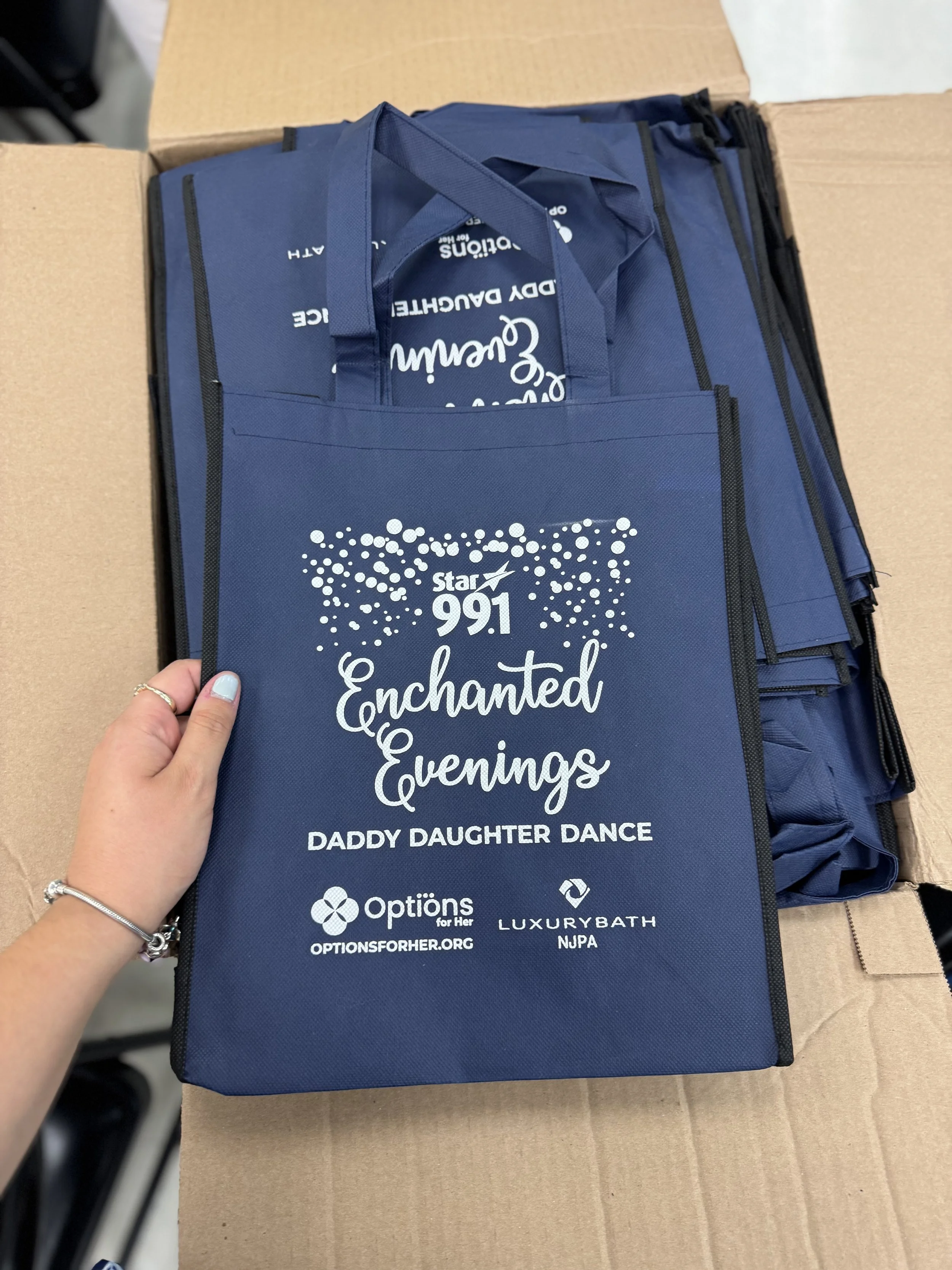 Blue tote bag with white text and logos, held by a person, on top of a cardboard box.