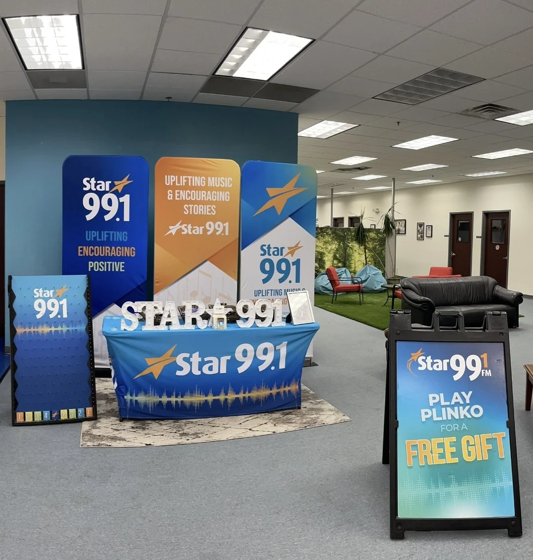 Indoor promotional display for Star 99.1 FM with banners, signs, and a table featuring branding and messages about uplifting music, encouraging stories, and a contest to play Plinko for a free gift. The background includes seating and decor in a loun