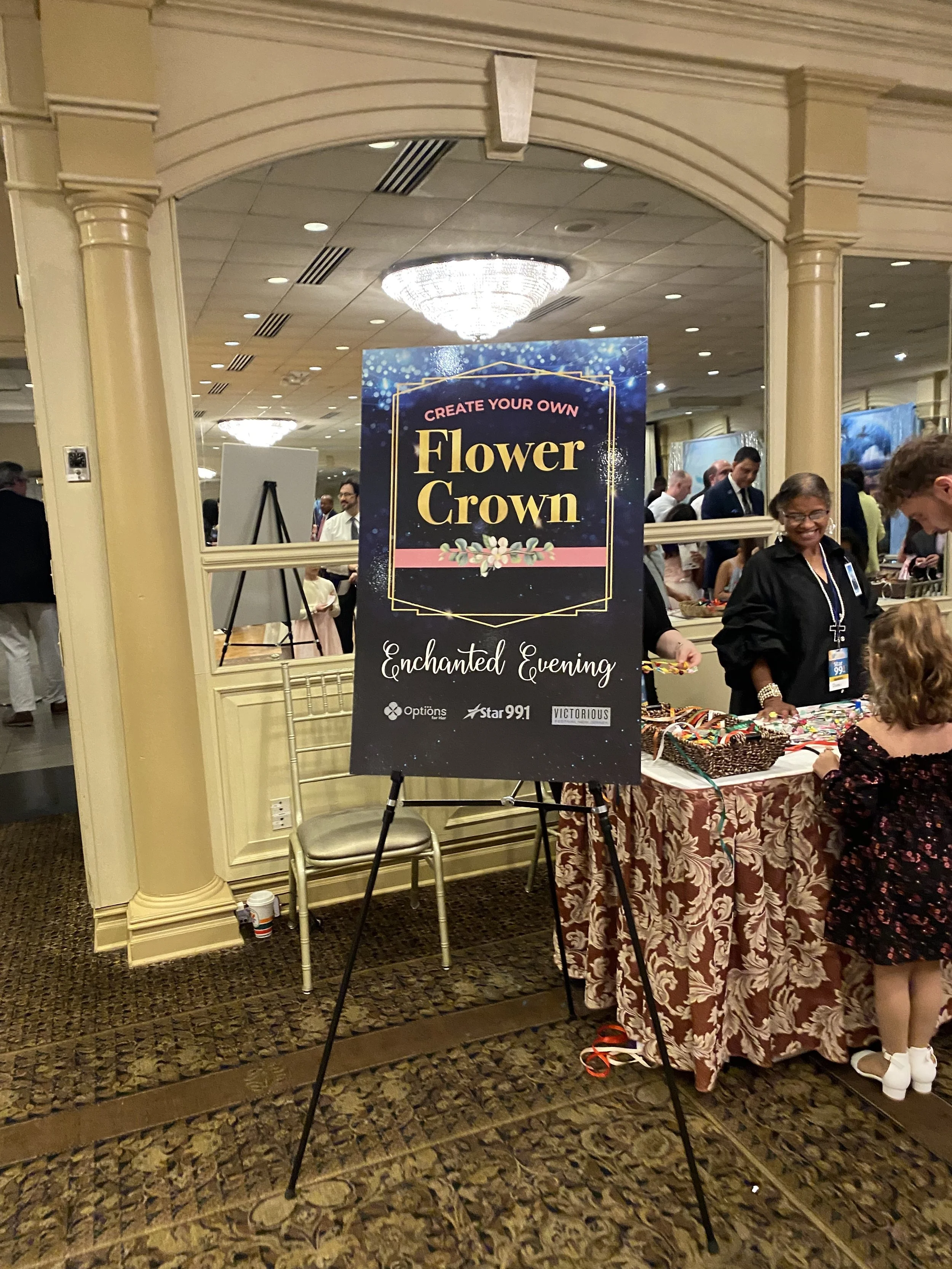 Signboard at an event titled 'Create Your Own Flower Crown, Enchanted Evening' with a decorated table handling flower crown making, surrounded by attendees, in an elegant banquet hall with chandeliers.
