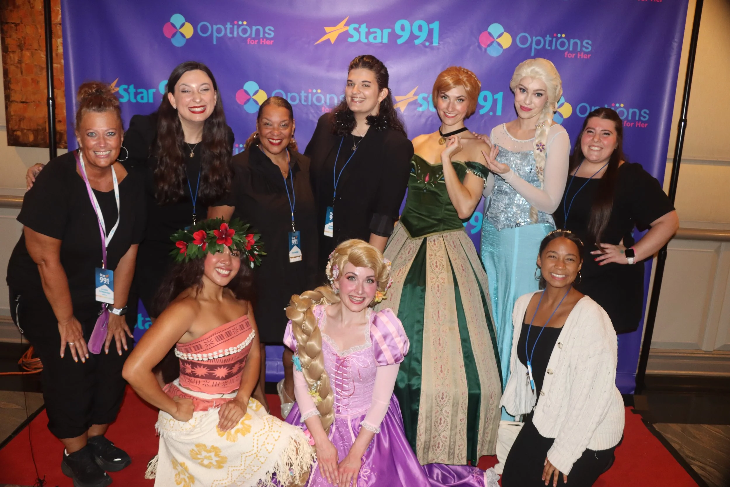 Group photo of women, including some dressed as Disney princess characters, standing and kneeling in front of a purple backdrop with logos for 'Options for Her' and 'Star 99.1' at an event.