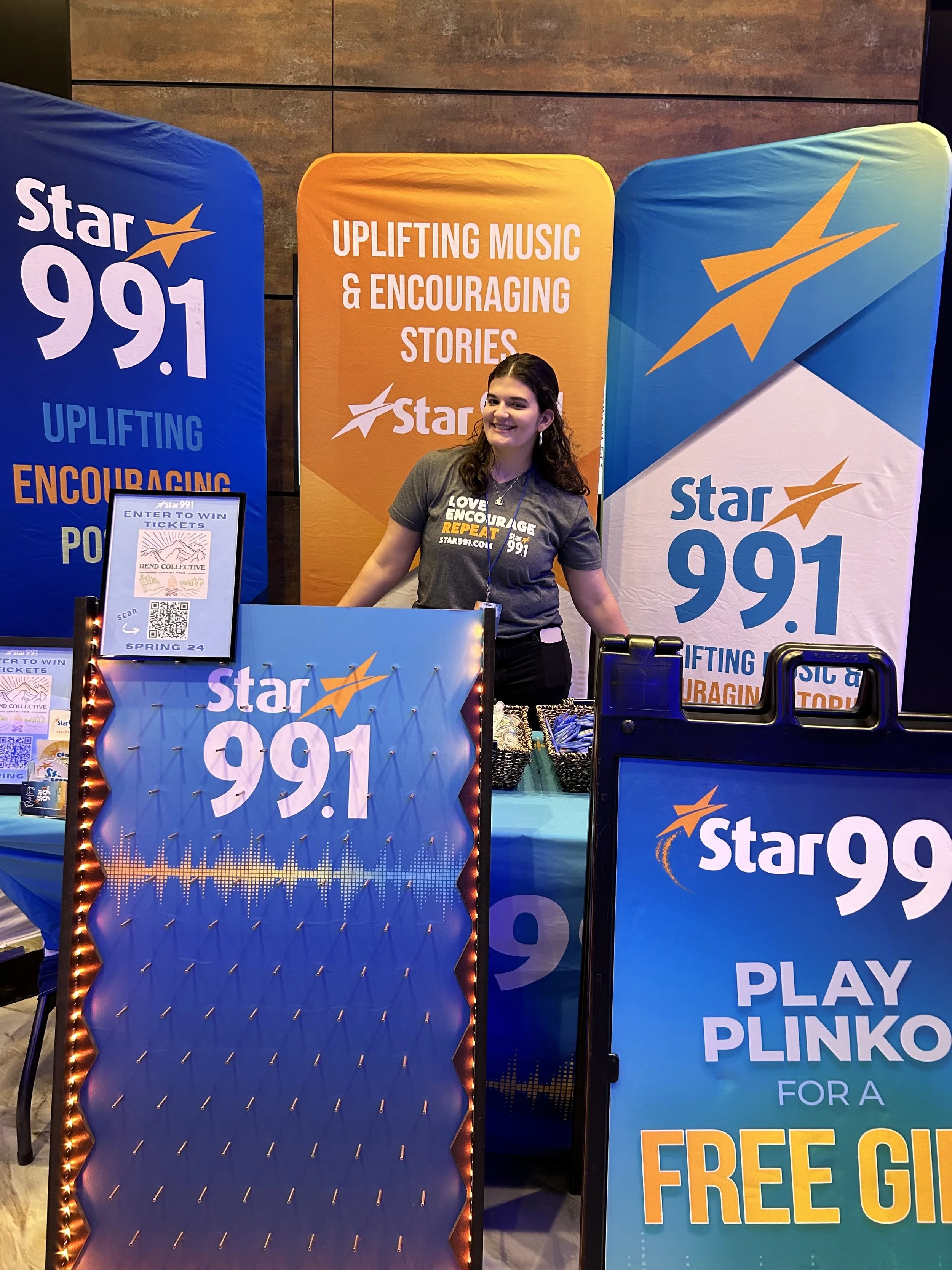 A woman standing at a promotional booth for Star 99.1 radio station, with banners and signs promoting the station and a giveaway. The woman is smiling and wearing a dark gray T-shirt with the station's logo.