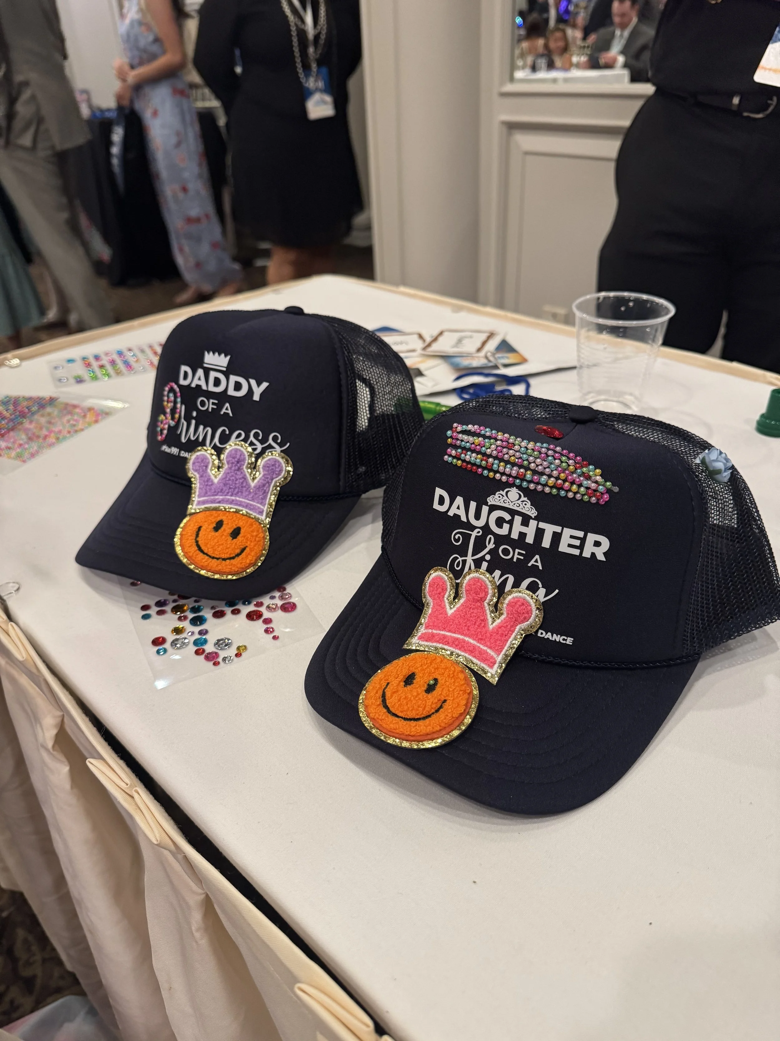 Black mesh trucker hats on a table with colorful jeweled embellishments and patches that say 'Daddy of a Princess' and 'Daughter of a King,' decorated with pink and purple crowns and orange smiley faces, at a busy indoor event.