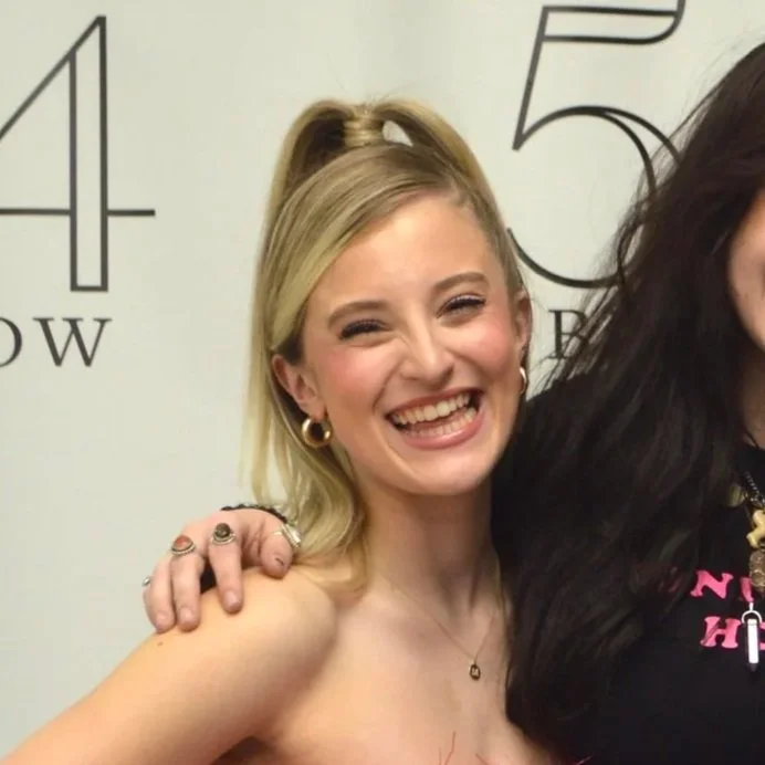 A young woman with blonde hair styled in a half-up, half-down hairstyle, smiling widely with her tongue out, wearing hoop earrings and a necklace, with her arm around another person.