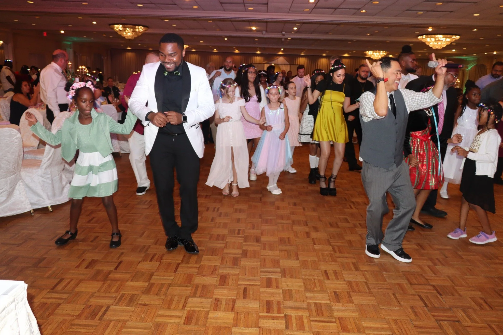 People dancing at an indoor celebration or party, including children and adults in formal and casual attire, with tables and chandeliers in the background.