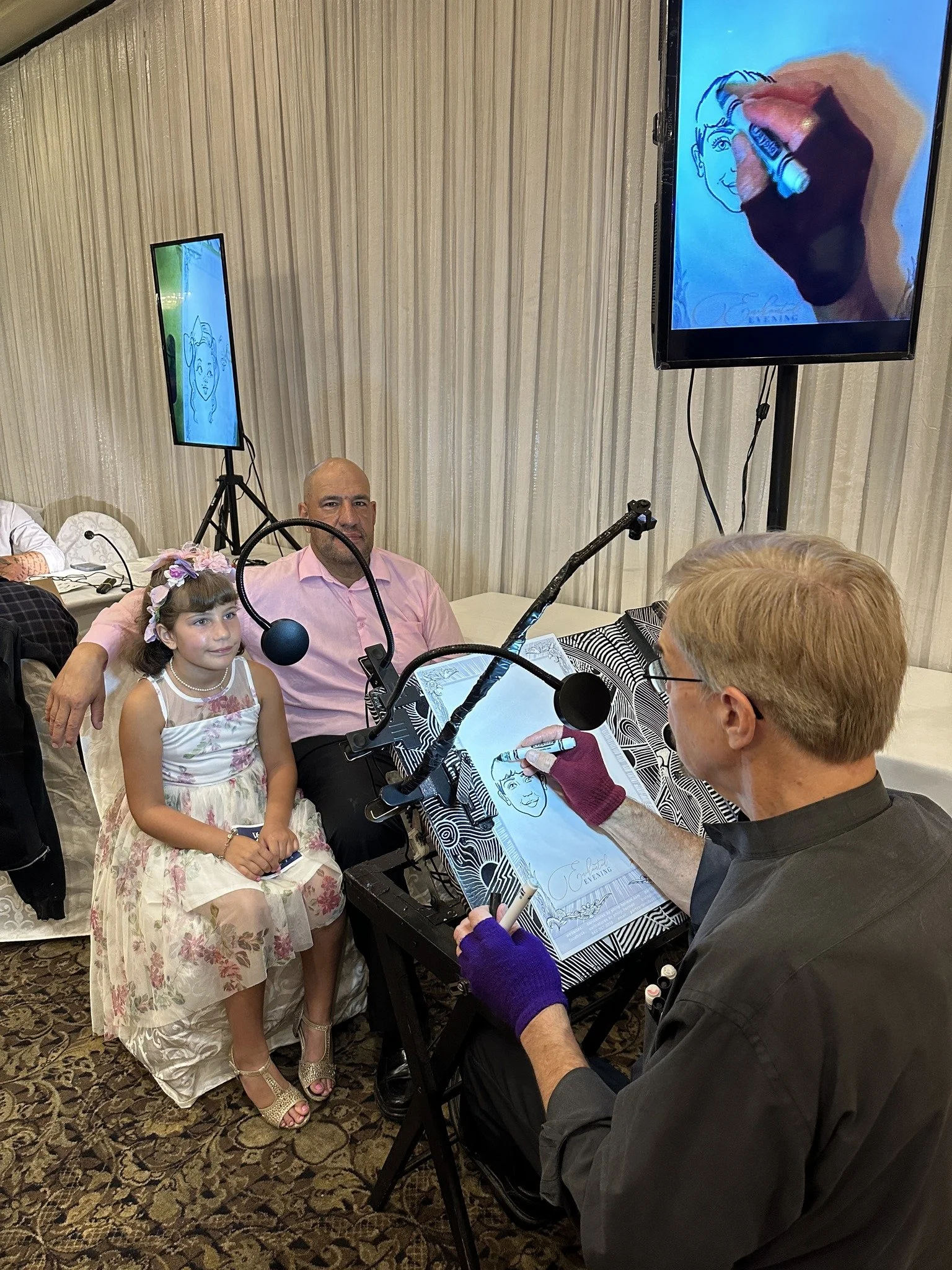 An artist is drawing a caricature of a little girl and a man at a table, with a monitor displaying the drawing process behind them in a decorated event space.