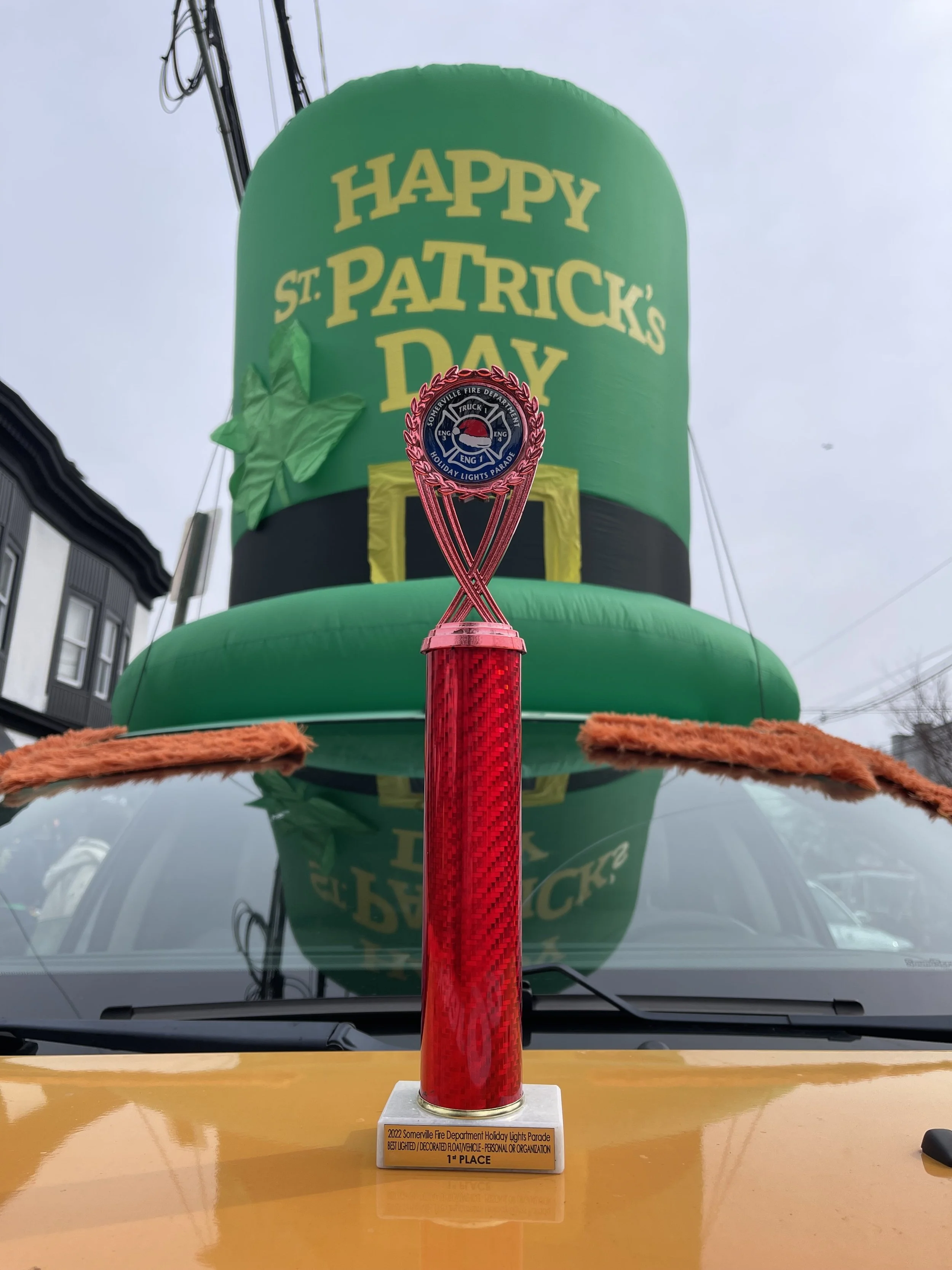 A red trophy with a black and blue emblem on the top, placed on a yellow vehicle, in front of a large green inflatable hat with yellow lettering that reads 'HAPPY ST. PATRICK'S DAY'.
