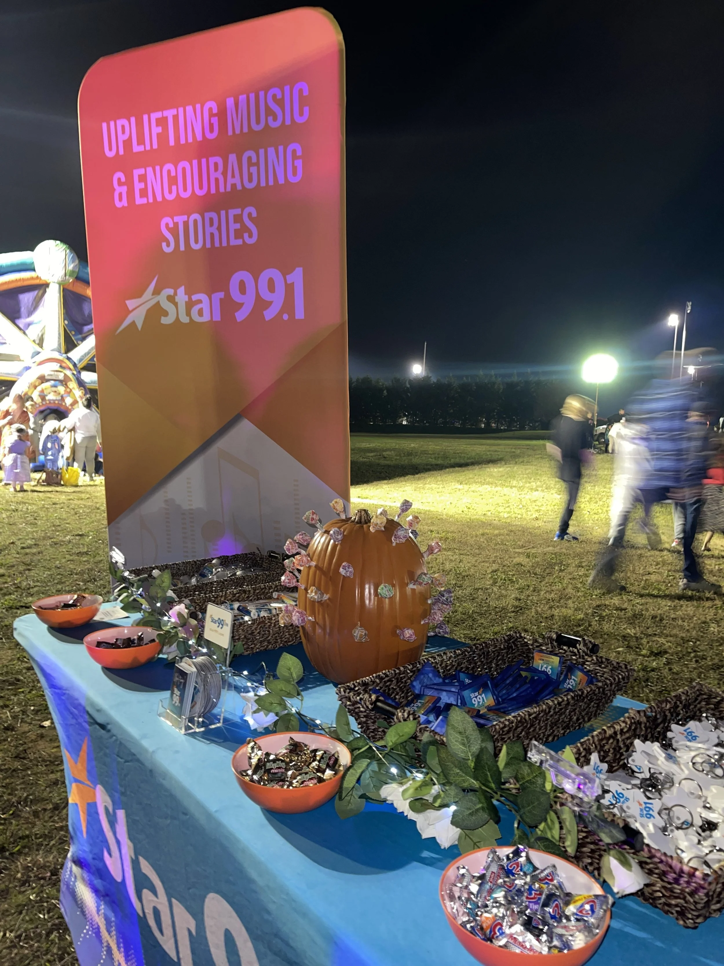 A booth with a sign reading 'Uplifting music & encouraging stories' for Star 99.1 radio station, set up at night with baskets of snacks and a pumpkin decoration, with people walking in the background.