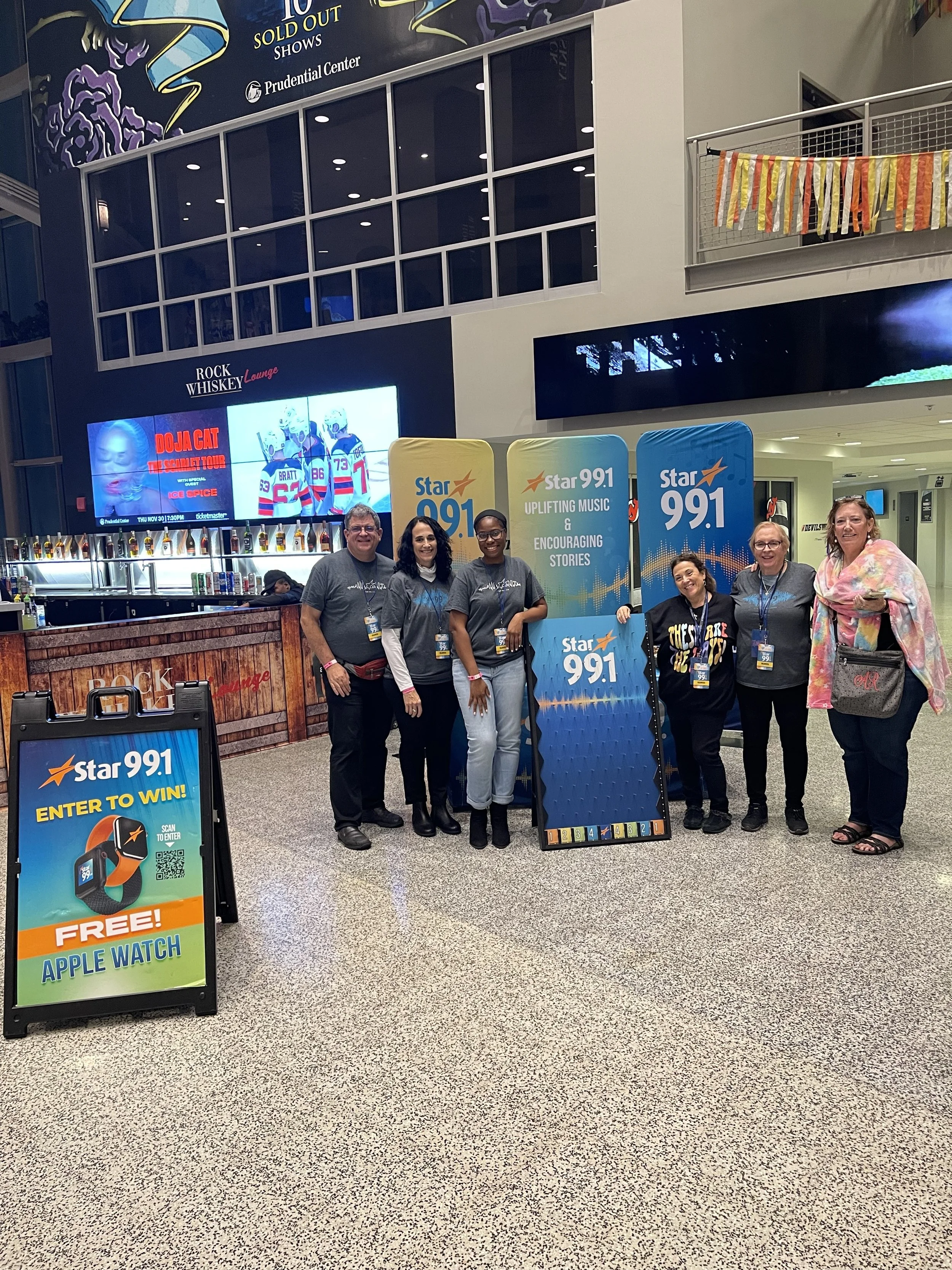 A group of six people standing together at a promotional event for Star 99.1 radio station, indoors at a venue with banners, digital screens, and a bar area in the background. They are smiling, wearing gray t-shirts, and holding promotional signs.