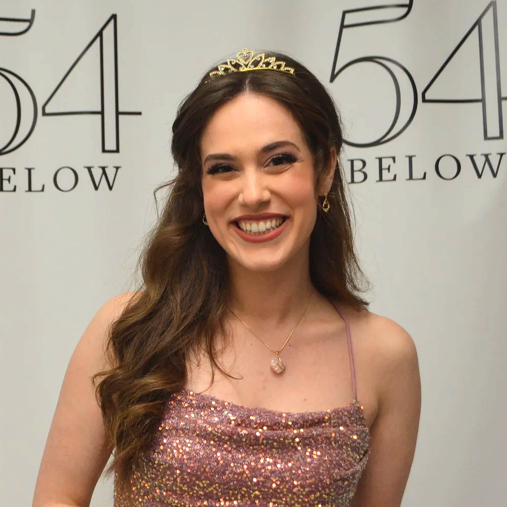 Young woman smiling, wearing a tiara and a sparkly pink dress, standing in front of a wall with large black numbers and the word 'BELOW'.