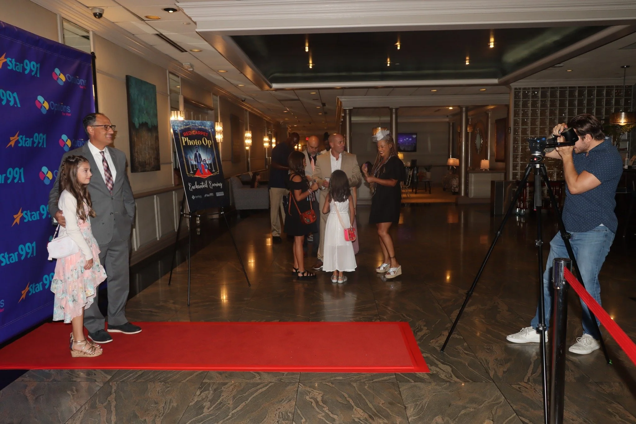 People attending a photo op event at an indoor venue, with a photographer taking pictures and a group of children and adults posing on the red carpet near a Star 99.1 backdrop and event signage.