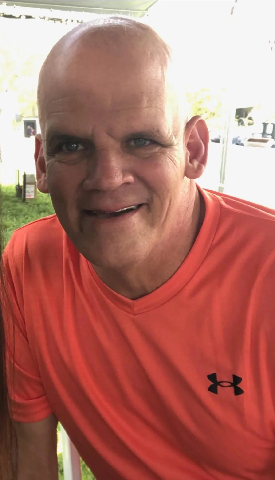 A man with a shaved head and light skin wearing an orange Under Armour shirt, smiling, in a bright outdoor setting.