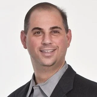 A headshot of a smiling man with short hair, wearing a gray shirt and a dark blazer, against a plain white background.