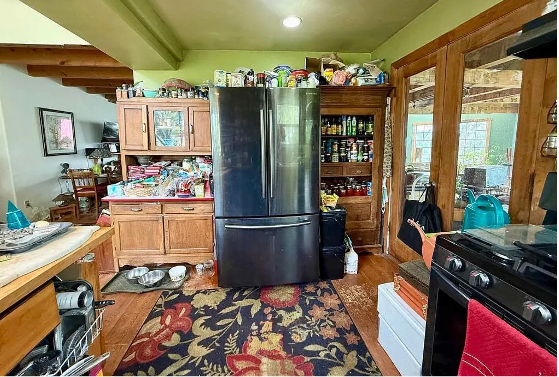 Cluttered rustic kitchen with black refrigerator, wooden cabinets, stocked pantry, black stove, and patterned floral rug