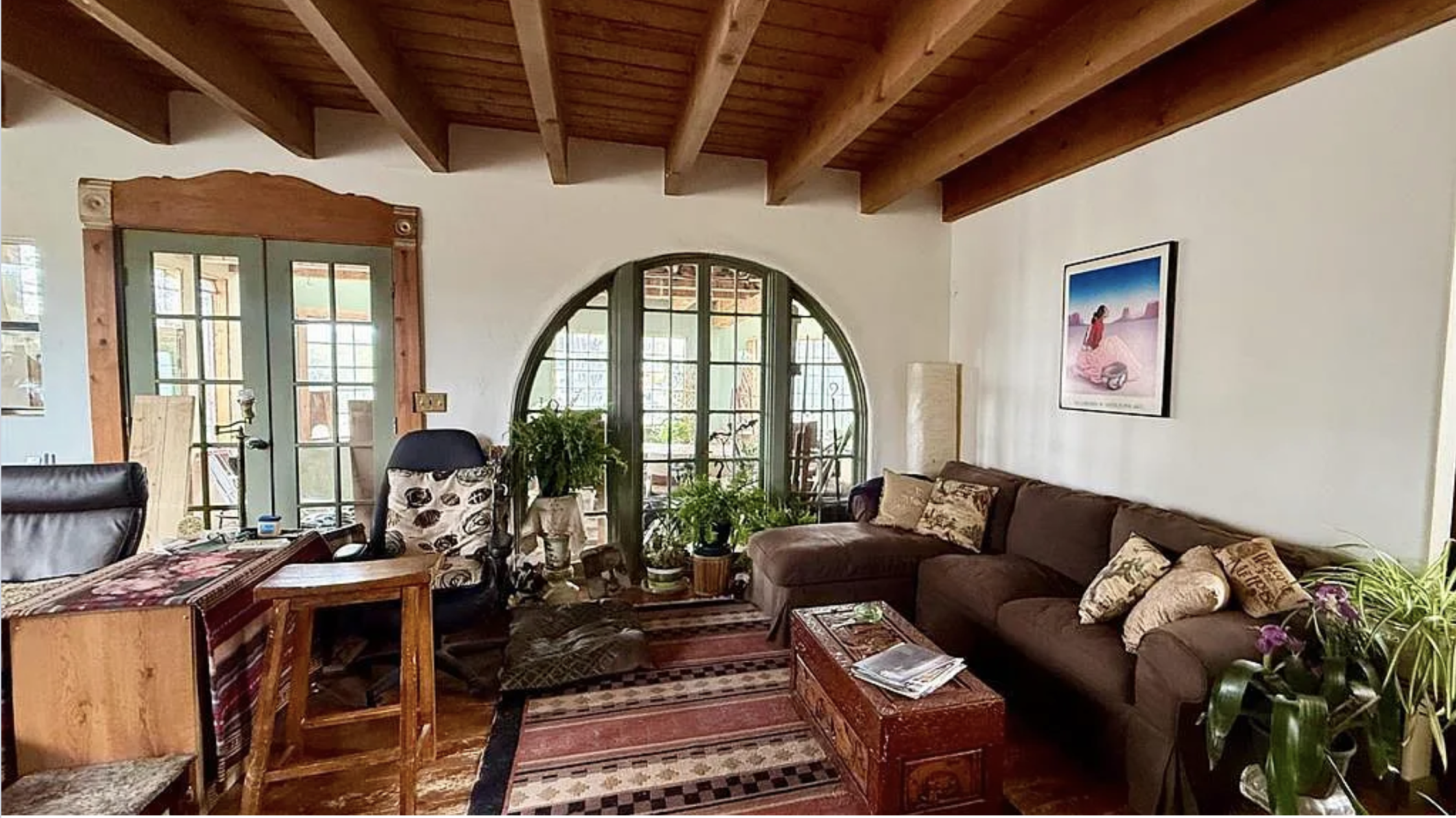 Rustic living room with exposed wood ceiling beams, arched windows, brown sectional sofa, plants, patterned rugs, and wooden coffee table