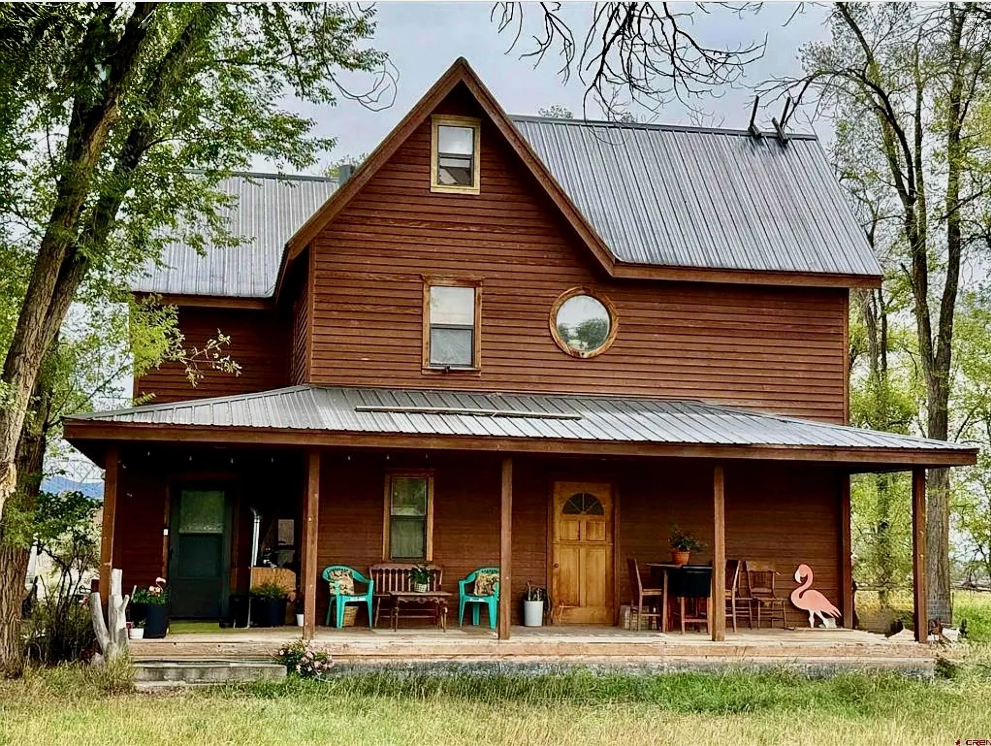 Rustic two-story wooden farmhouse with a metal roof, wraparound porch, outdoor seating, and pink flamingo decoration surrounded by trees