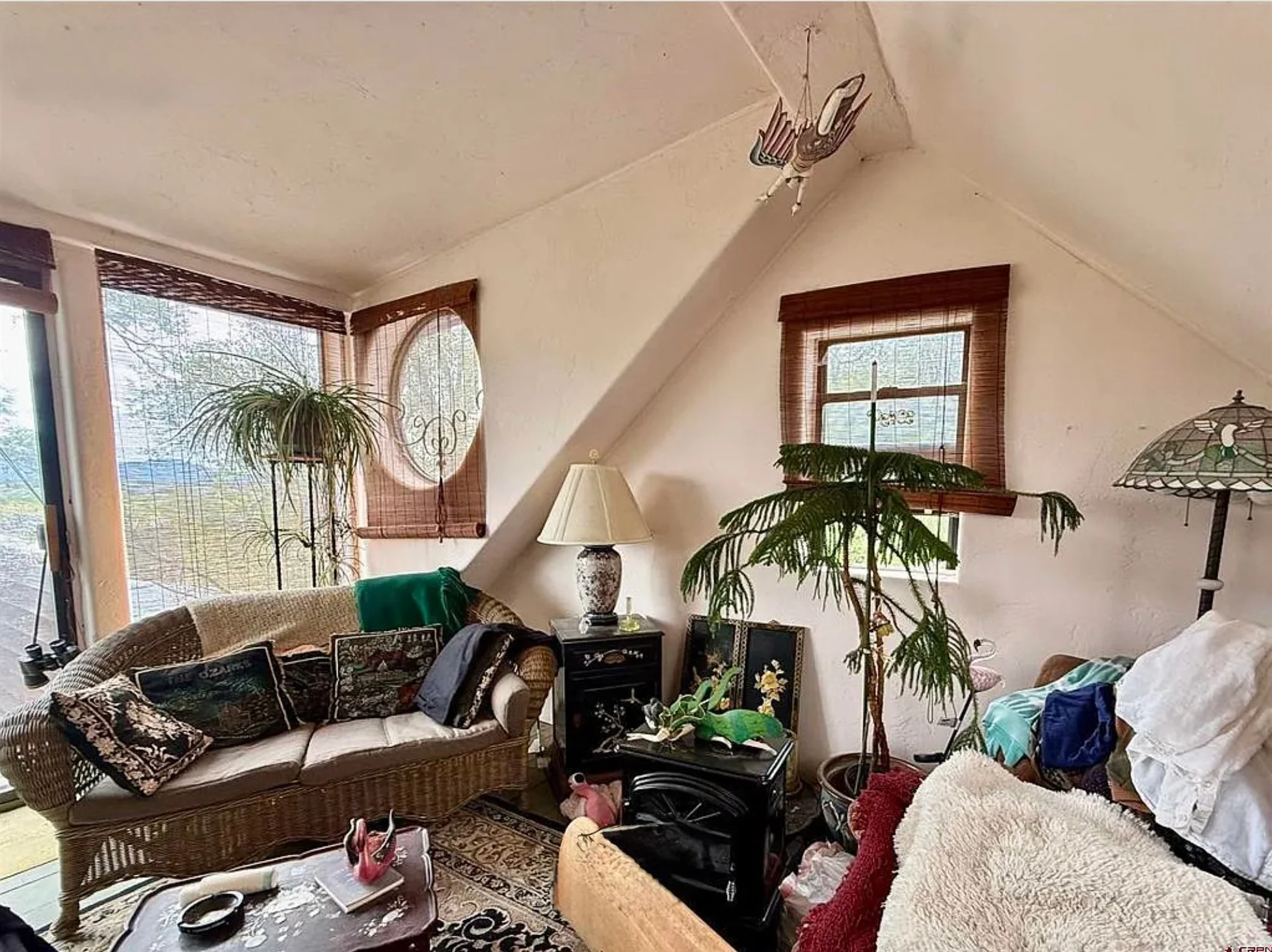 Cozy living room with wicker sofa, houseplants, bamboo shades, decorative lamps, and a fireplace.