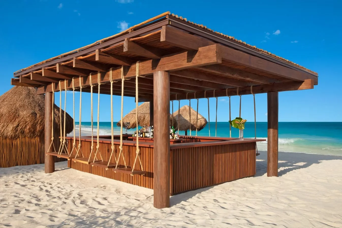 Beachside bar with wooden structure, swing seats, and thatched umbrellas, overlooking ocean with blue skies.