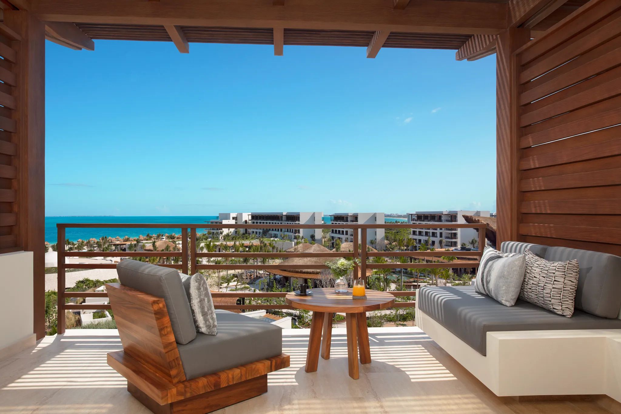 Balcony seating area with two cushioned chairs and a small round wooden table overlooking a coastal view with modern white buildings, palm trees, and the ocean under a clear blue sky.