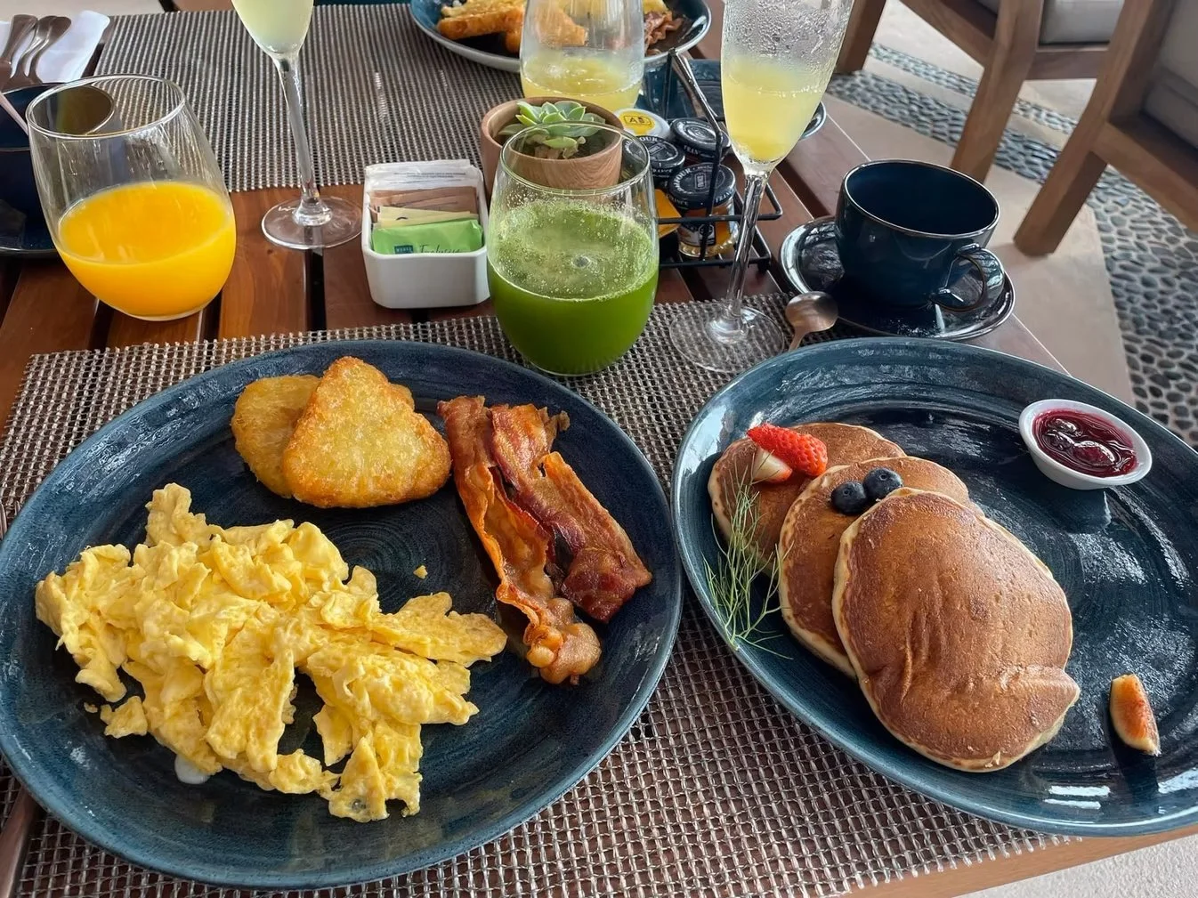 Breakfast spread includes scrambled eggs, bacon, hash browns, pancakes with berries and syrup, and beverages such as orange juice, green juice, coffee, and champagne on a table with placemats.