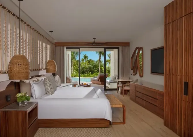 Hotel room with two beds, wooden furniture, a flat-screen TV, and a balcony view of palm trees and a swimming pool outside.