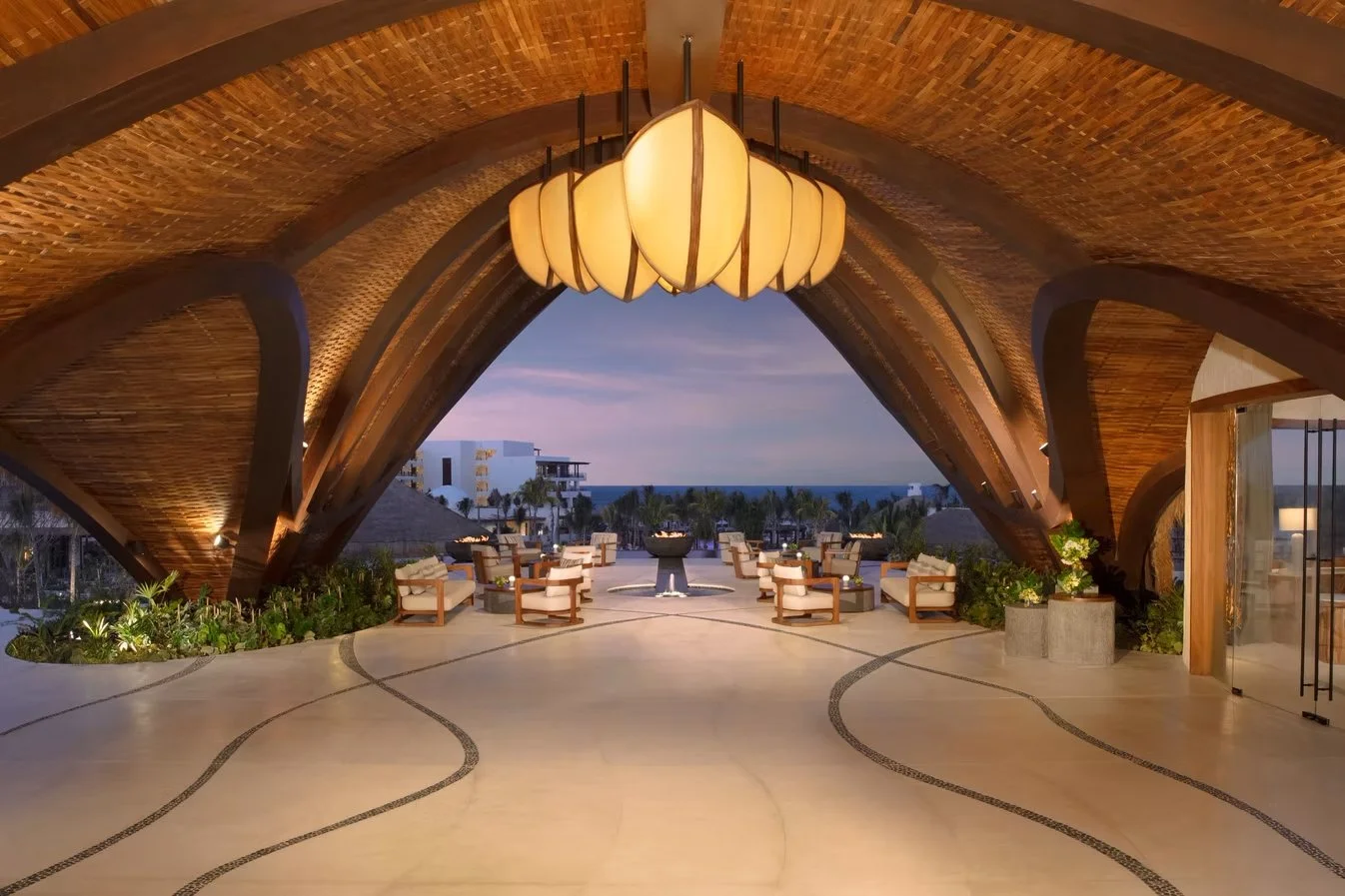 Luxury outdoor terrace with a large wooden vaulted ceiling, a central chandelier, and seating with armchairs and small tables, overlooking a scenic view of the ocean and skyline at sunset.