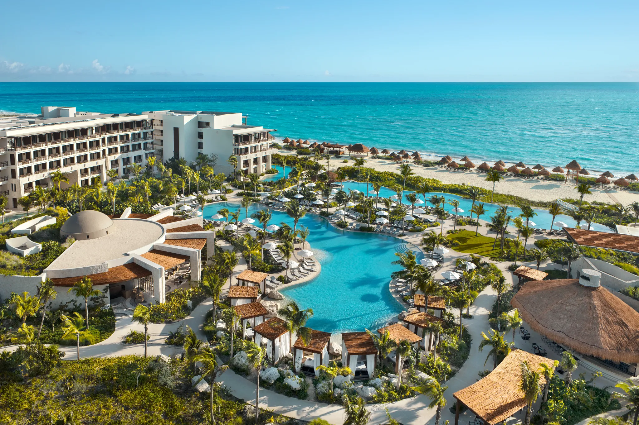 Aerial view of a luxury beachfront resort with multiple swimming pools, palm trees, and sandy beach with thatched umbrellas facing the ocean under a clear blue sky.