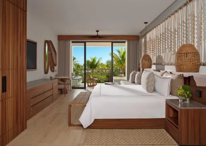 Modern hotel room with a large bed, decorative pillows, wooden furniture, and a balcony view of palm trees and clear skies.