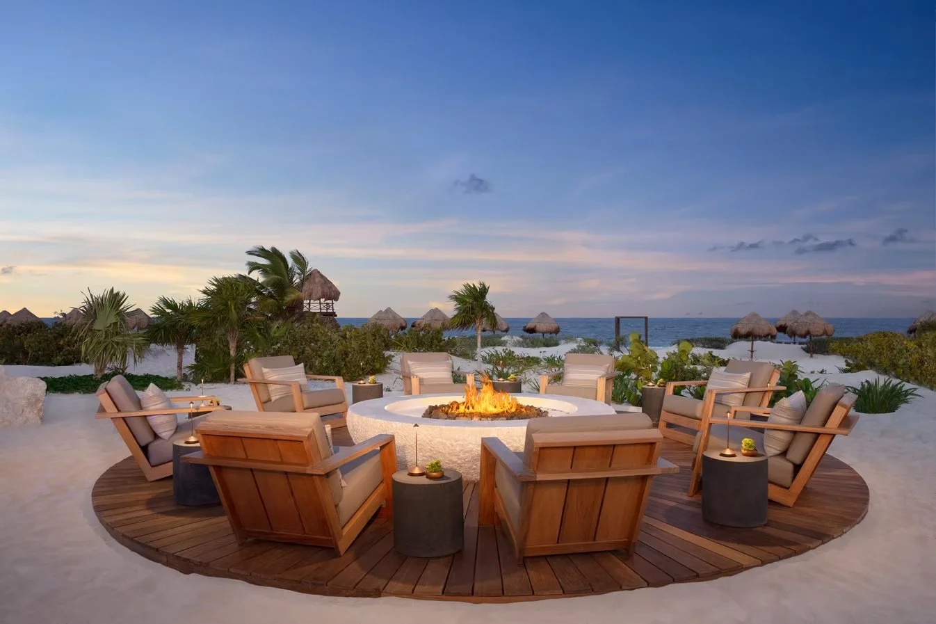 Beachside outdoor seating area with a fire pit, surrounded by wooden chairs, and thatched umbrellas in the background, with an ocean view at sunset.