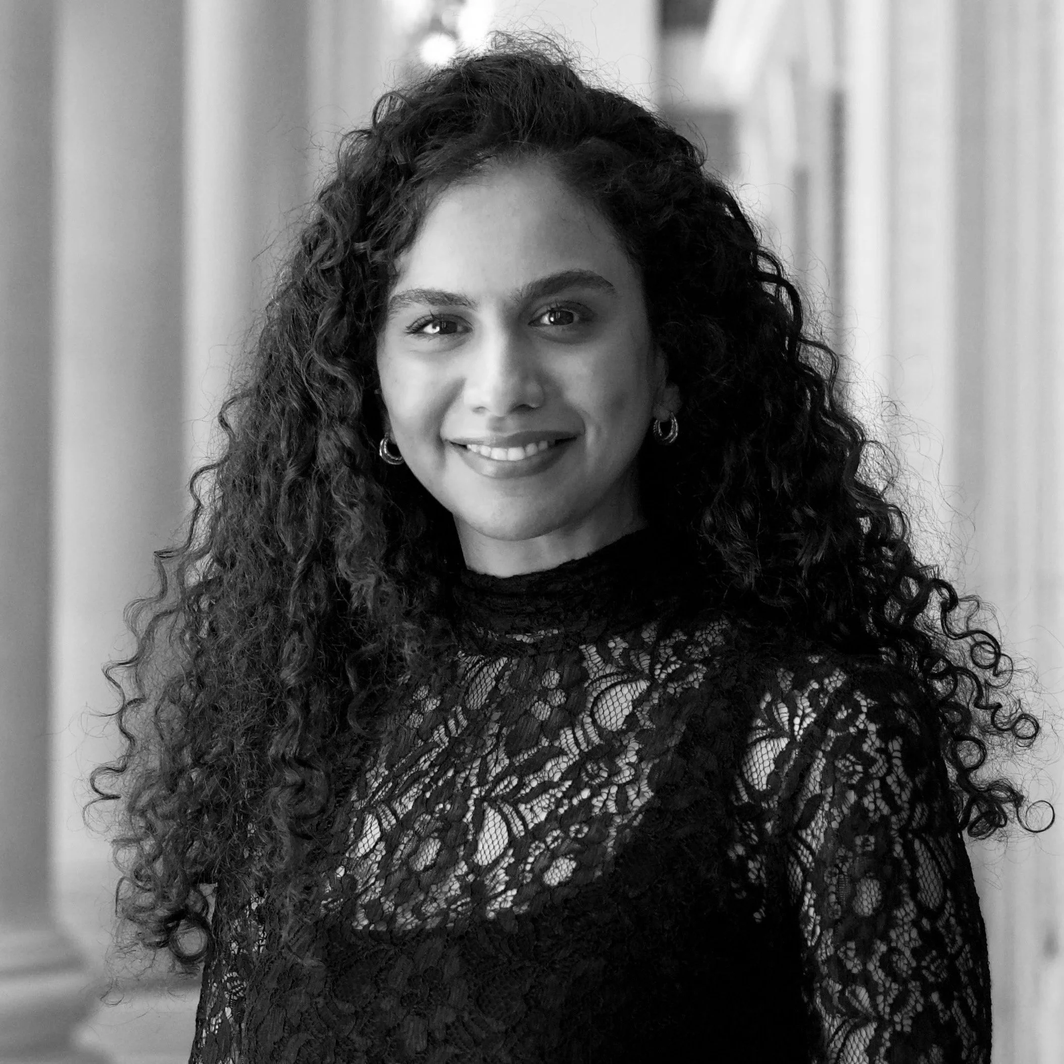 A black and white portrait of a woman with long curly hair, wearing a lace top and hoop earrings, standing in front of a blurred background.