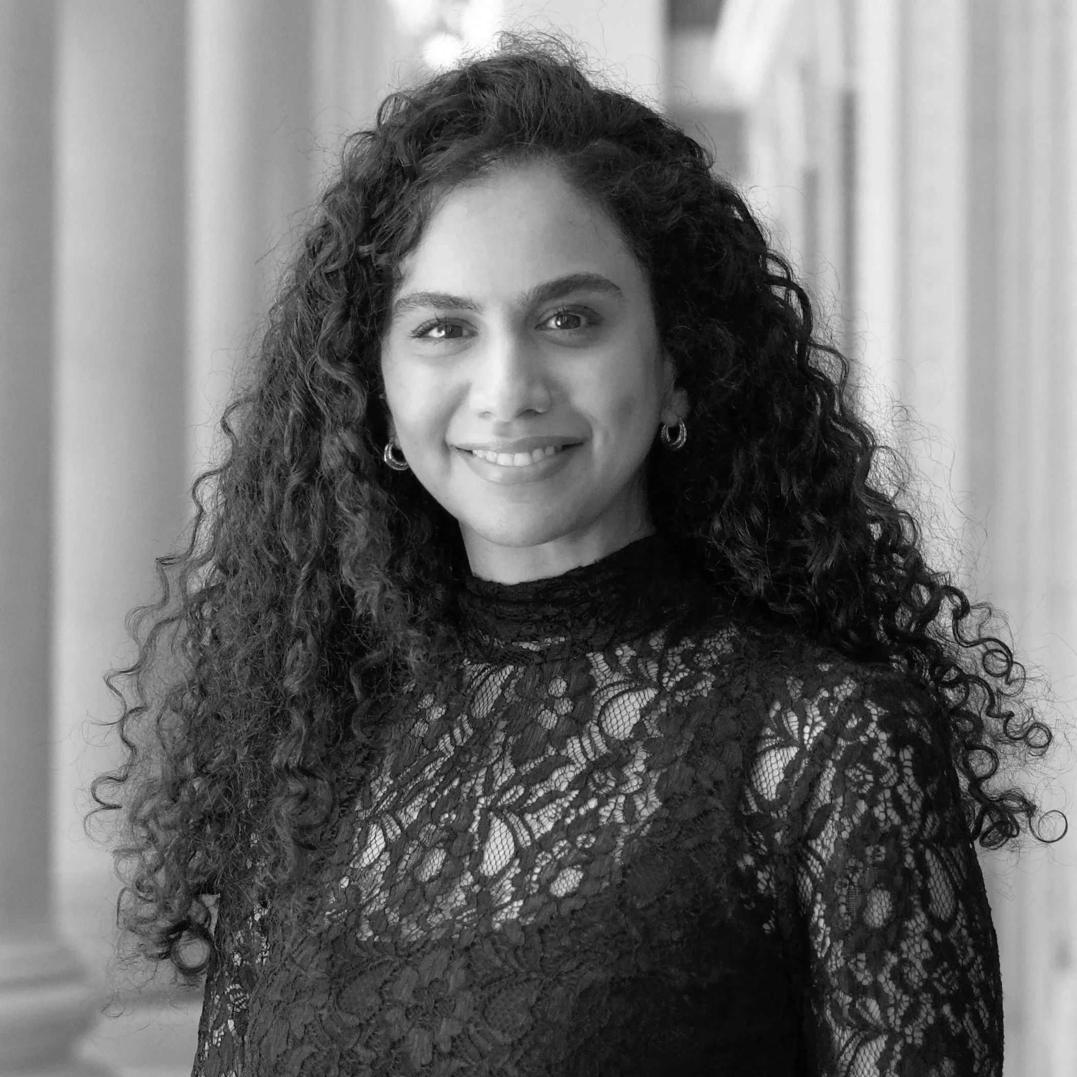 Portrait of a woman smiling, wearing a lace top, with curly hair, earrings, in a setting with columns.
