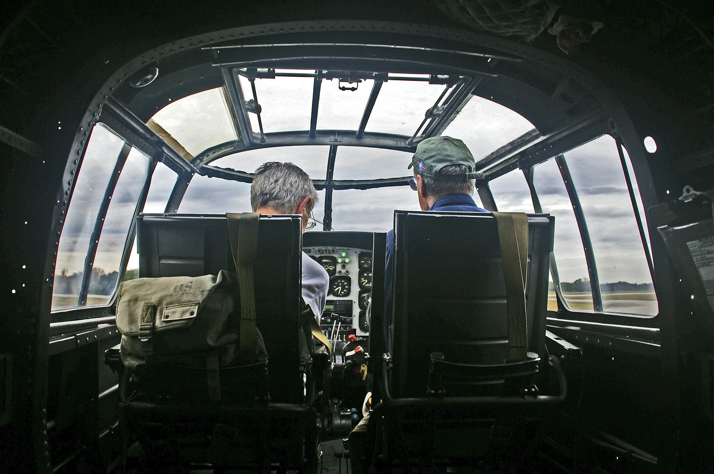 View from inside a vintage aircraft cockpit showing two pilots operating the controls during daytime, with a cloudy sky outside.
