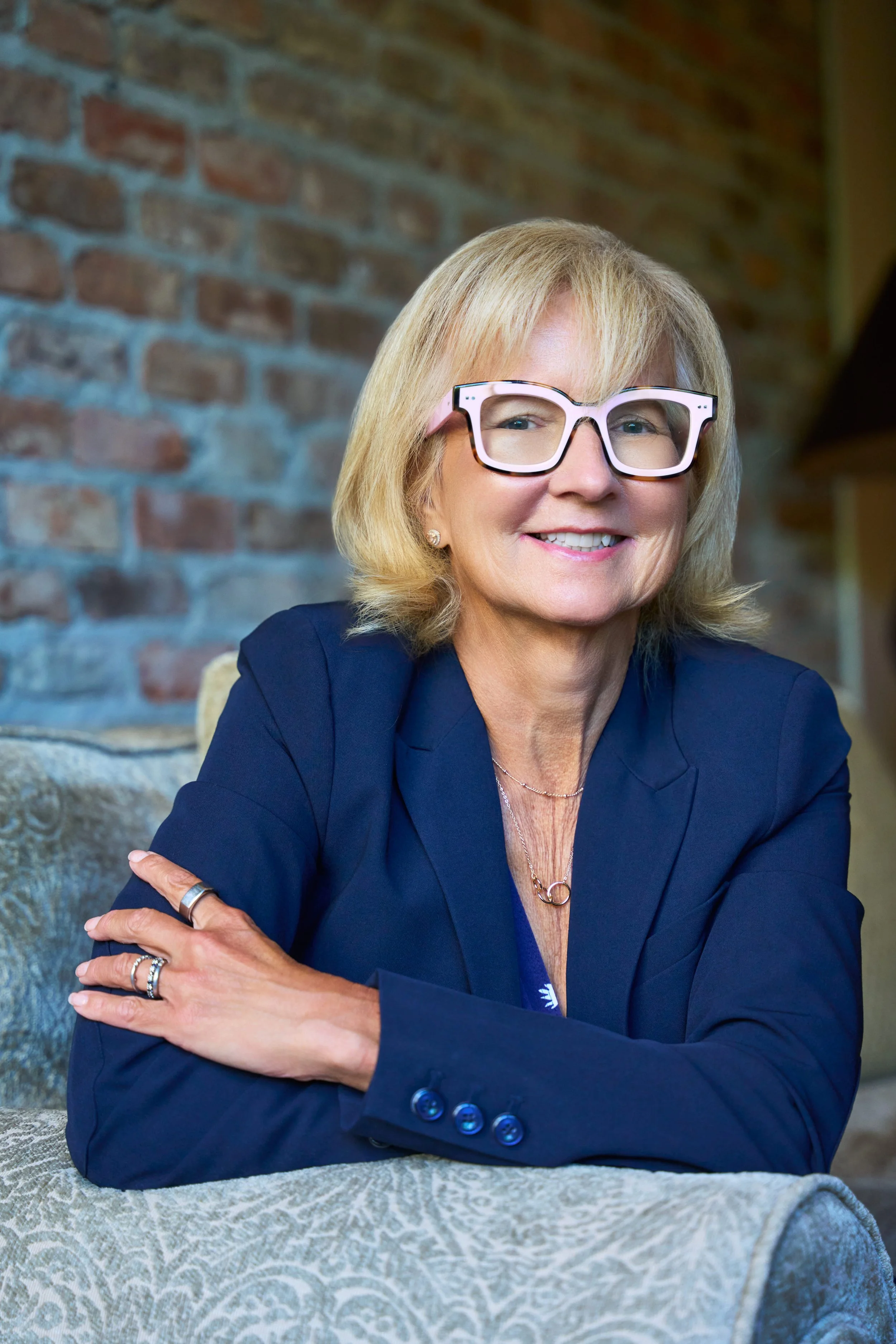 A smiling woman with shoulder-length blonde hair, wearing large white glasses, a navy blazer, and gold jewelry, sitting at a table with a patterned tablecloth against a brick wall background.