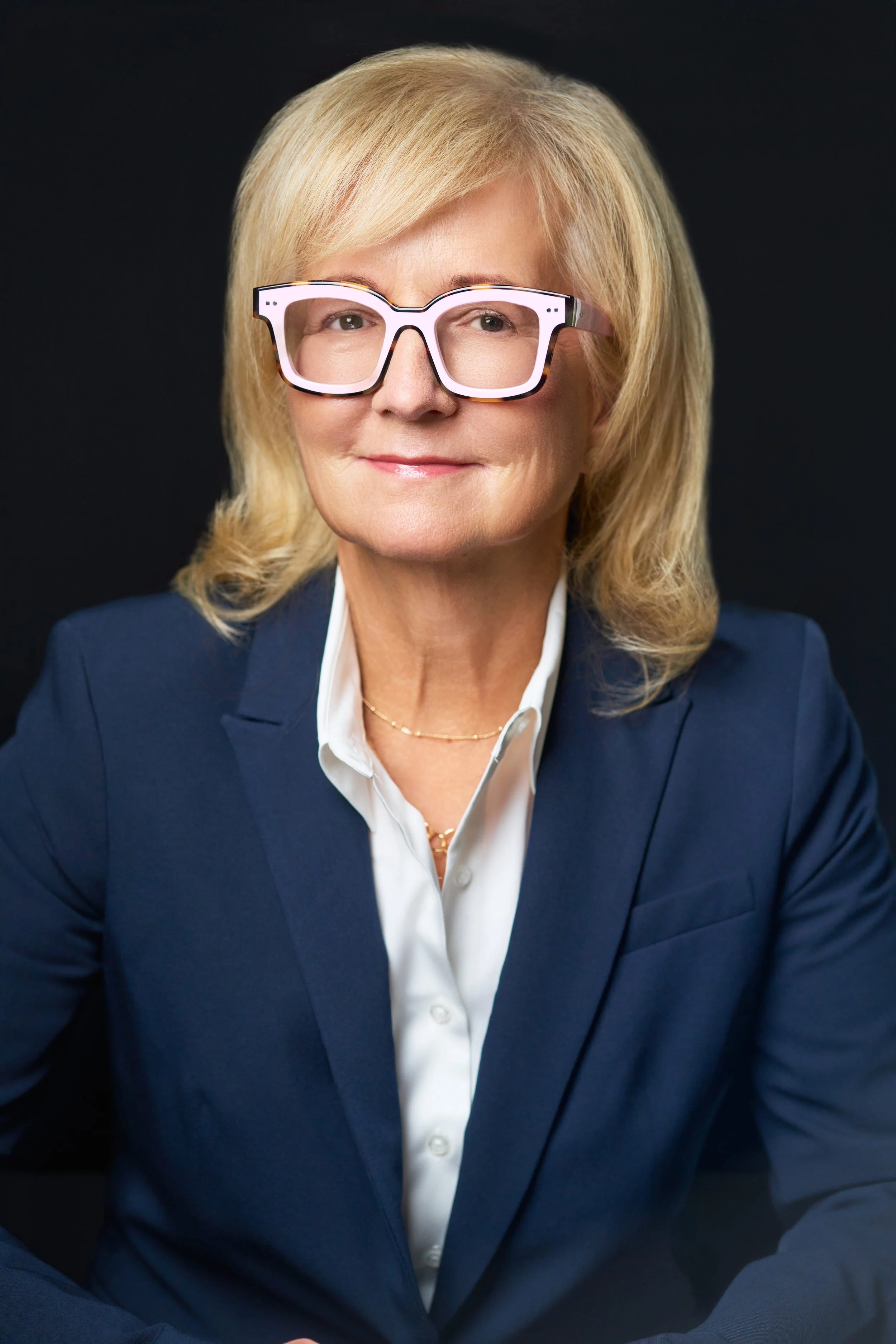 A woman in a dark blue blazer and white shirt, wearing large white and black glasses, sitting against a black background.