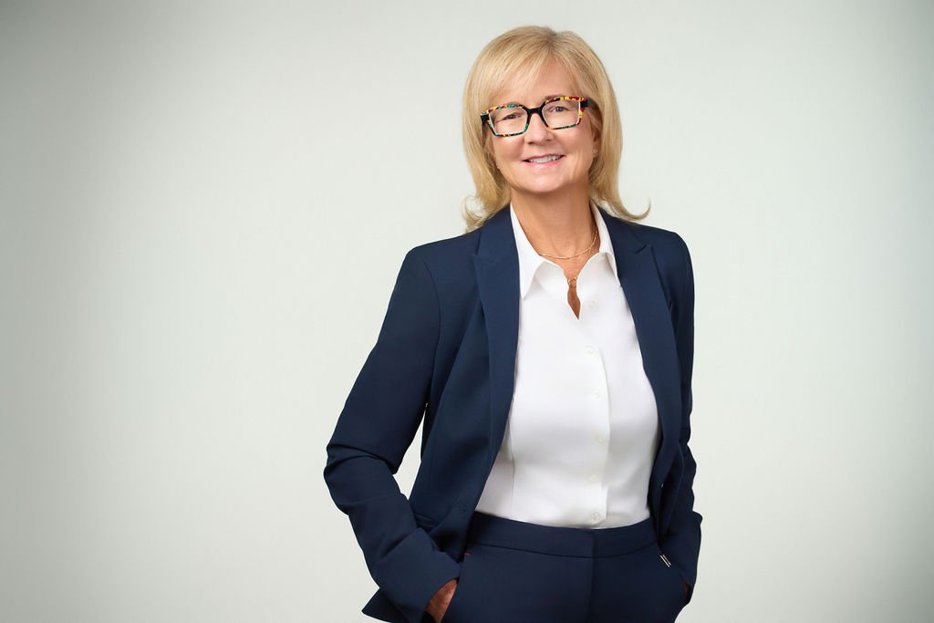 A middle-aged woman with blonde hair and glasses, dressed in a navy blazer and white shirt, smiling confidently with her hands in her pockets, standing against a plain light background.
