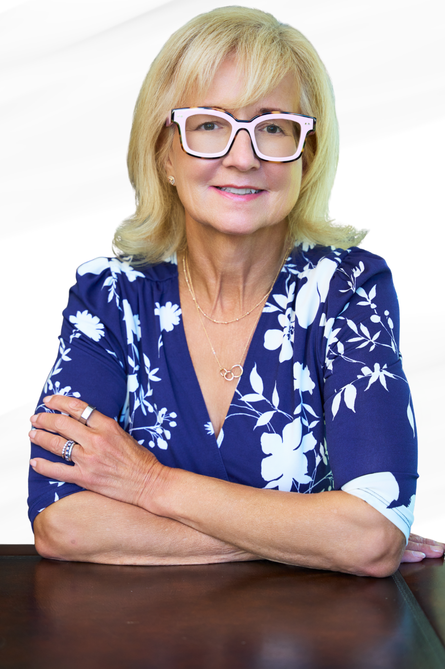 A blonde woman wearing glasses and a blue floral dress, sitting with her arms crossed on a dark wooden table.