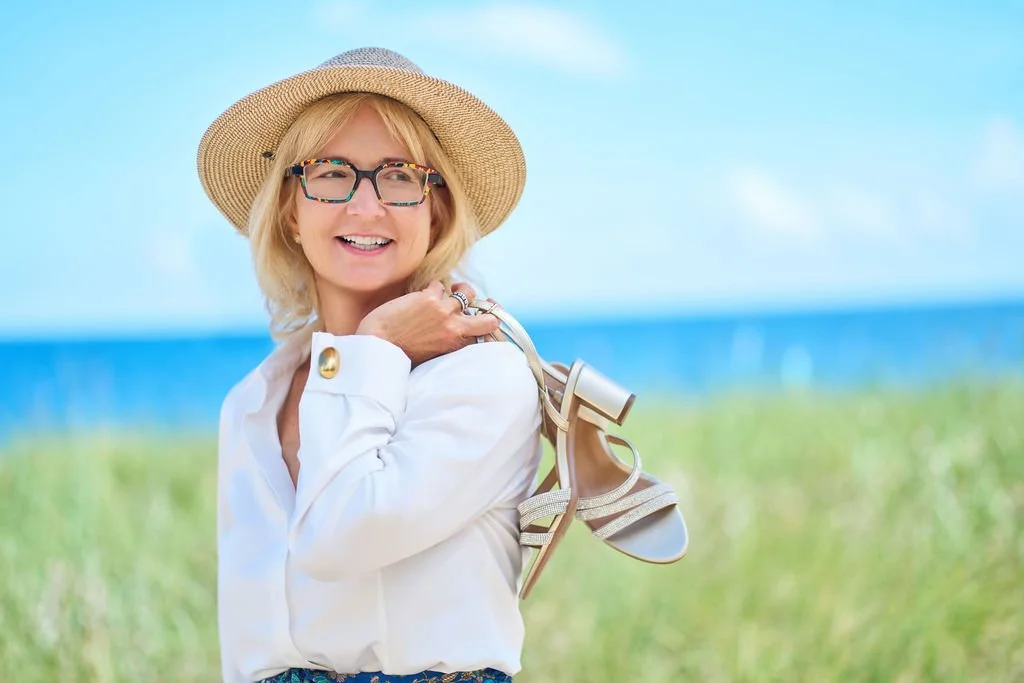 Smiling woman in a sunhat and glasses holding sandals at the beach.