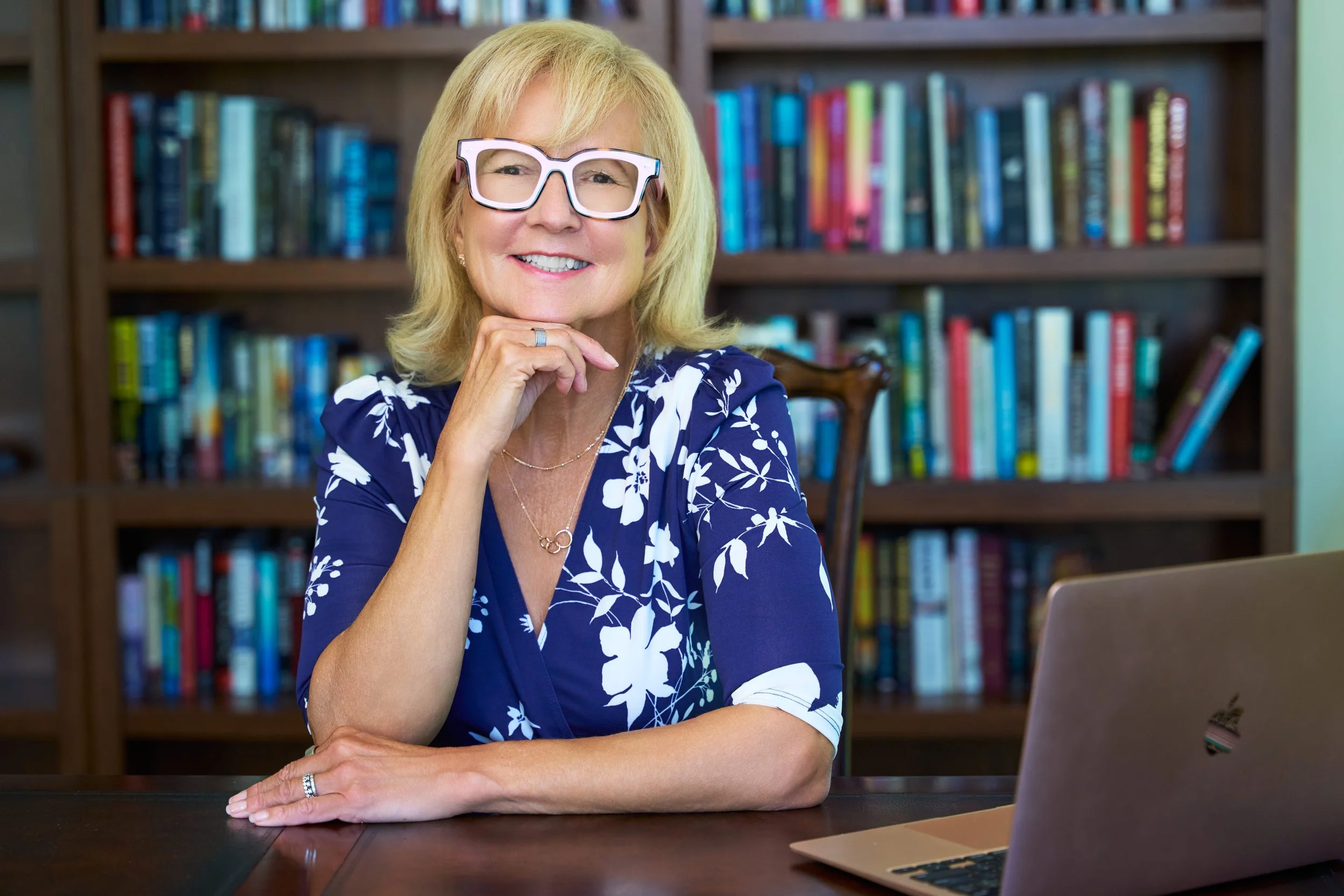 A smiling middle-aged woman with blonde hair and glasses, sitting at a dark wooden table with a laptop in front of her, in a room with a background of bookshelves filled with colorful books.