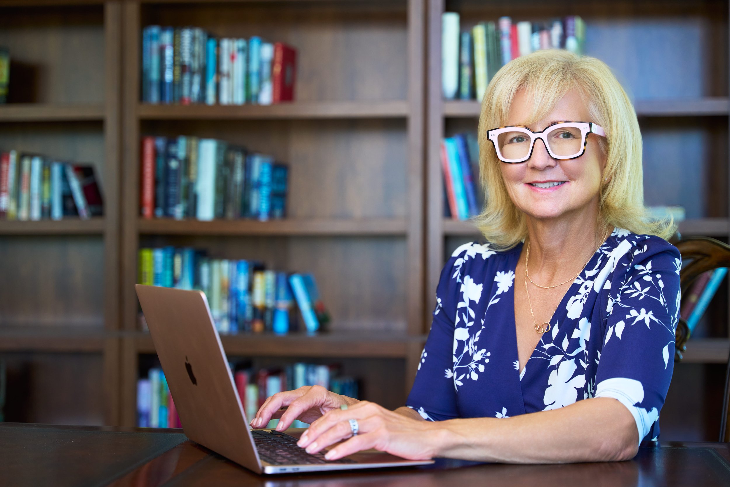 A woman with blonde hair wearing glasses and a blue and white floral dress working on a laptop at a wooden table with a background of bookshelves filled with colorful books.