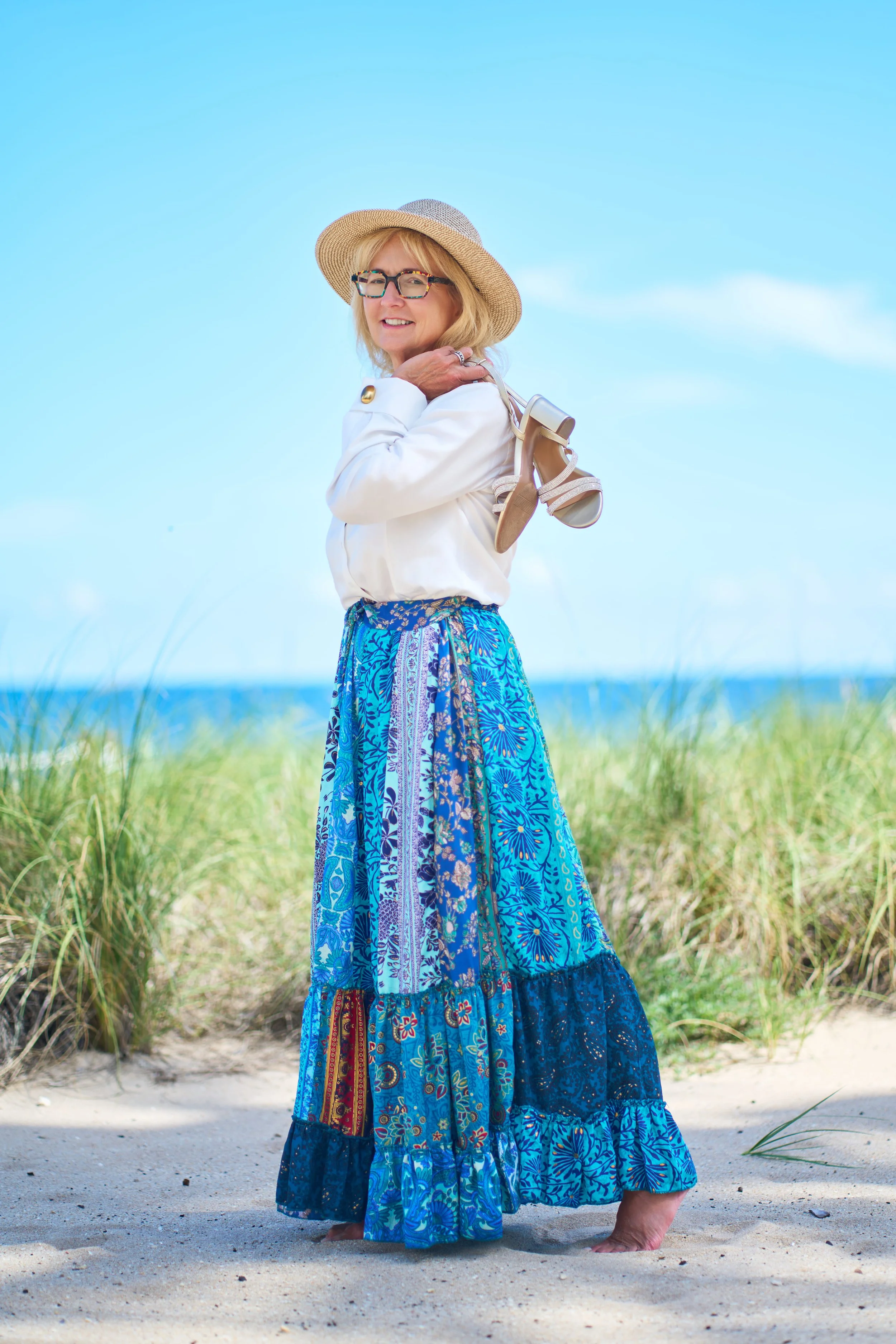 Woman in dress walking barefoot on beach, holding hat, with blue sky and ocean behind her.