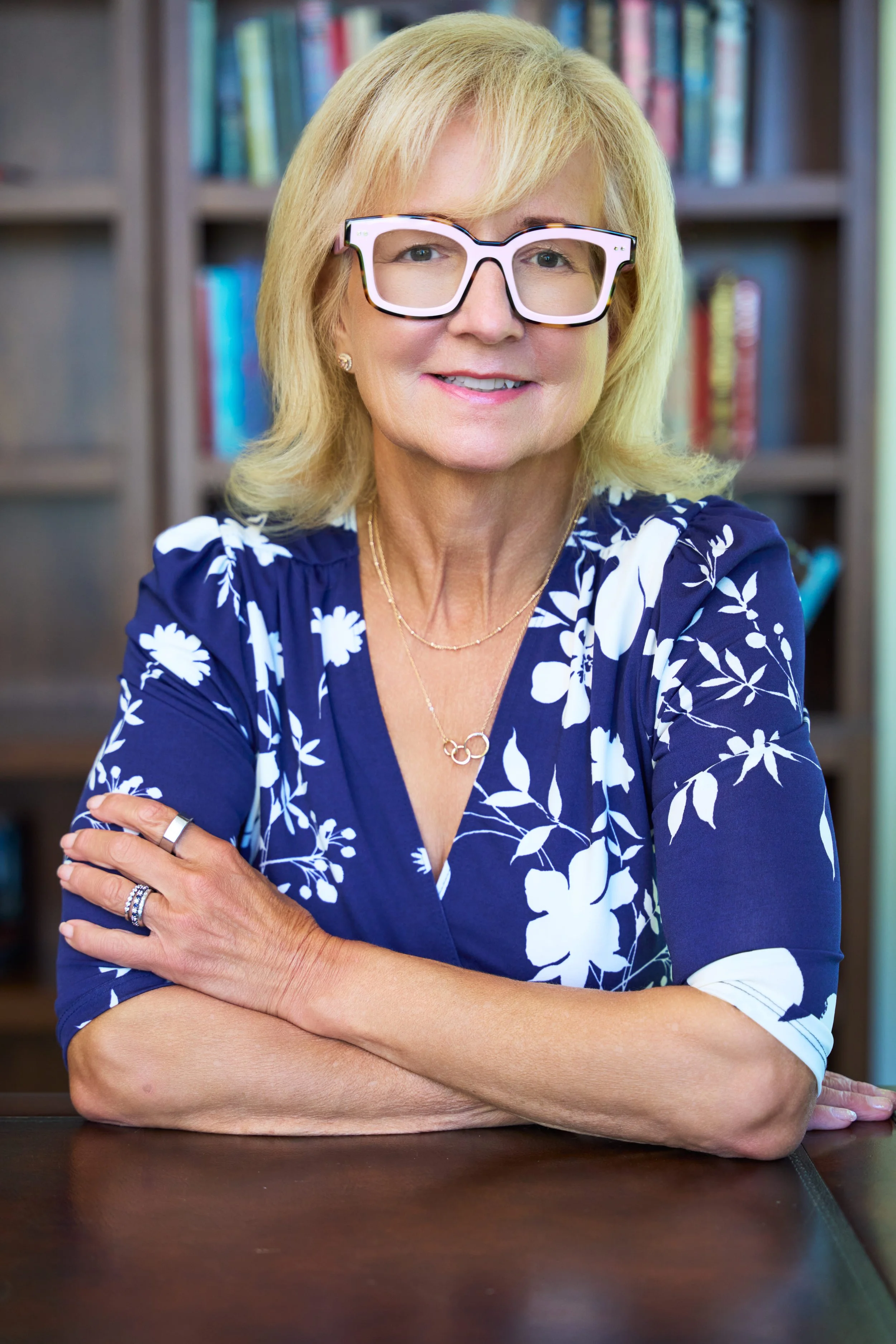 A woman with blonde hair wearing large white and black glasses, a blue and white floral dress, jewelry, and rings, sitting with arms crossed in front of a bookshelf filled with books.