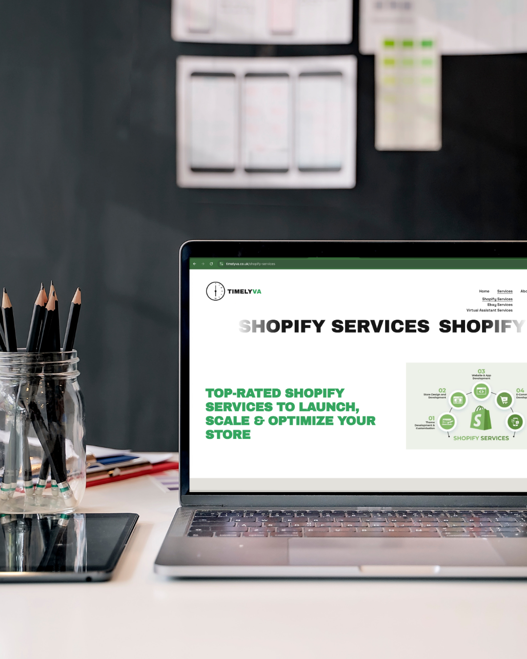 Laptop on a white desk displaying a website for Shopify services, with a glass jar filled with black pencils and a tablet nearby, against a dark wall with blurred charts or schedules on the wall.