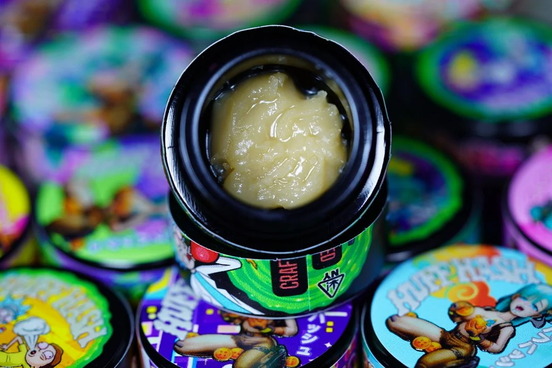 Open container of cannabis THCA concentrate surrounded by colorful containers with cartoon characters and vibrant designs.